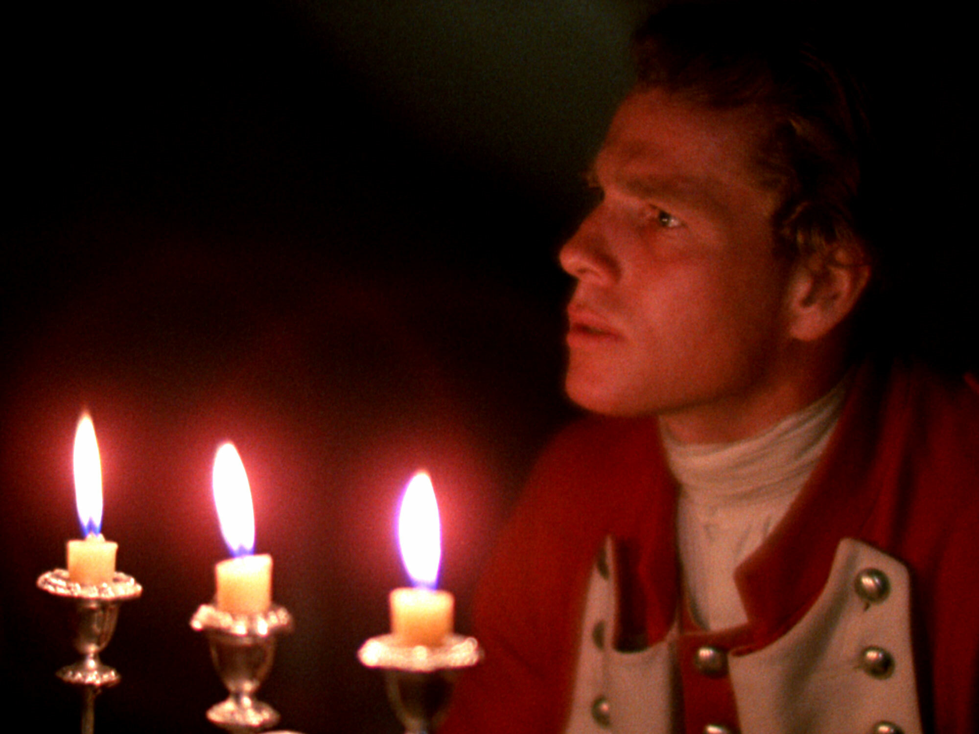 A person with curly hair sits in a dark room, illuminated by the glow of three lit candles.