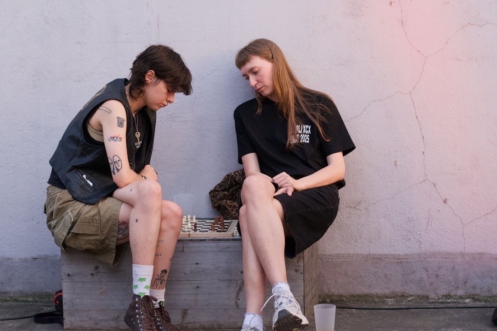 Two people sitting on concrete ledge playing chess. One wears dark vest and shorts, the other a black t-shirt. Pink wall behind them.