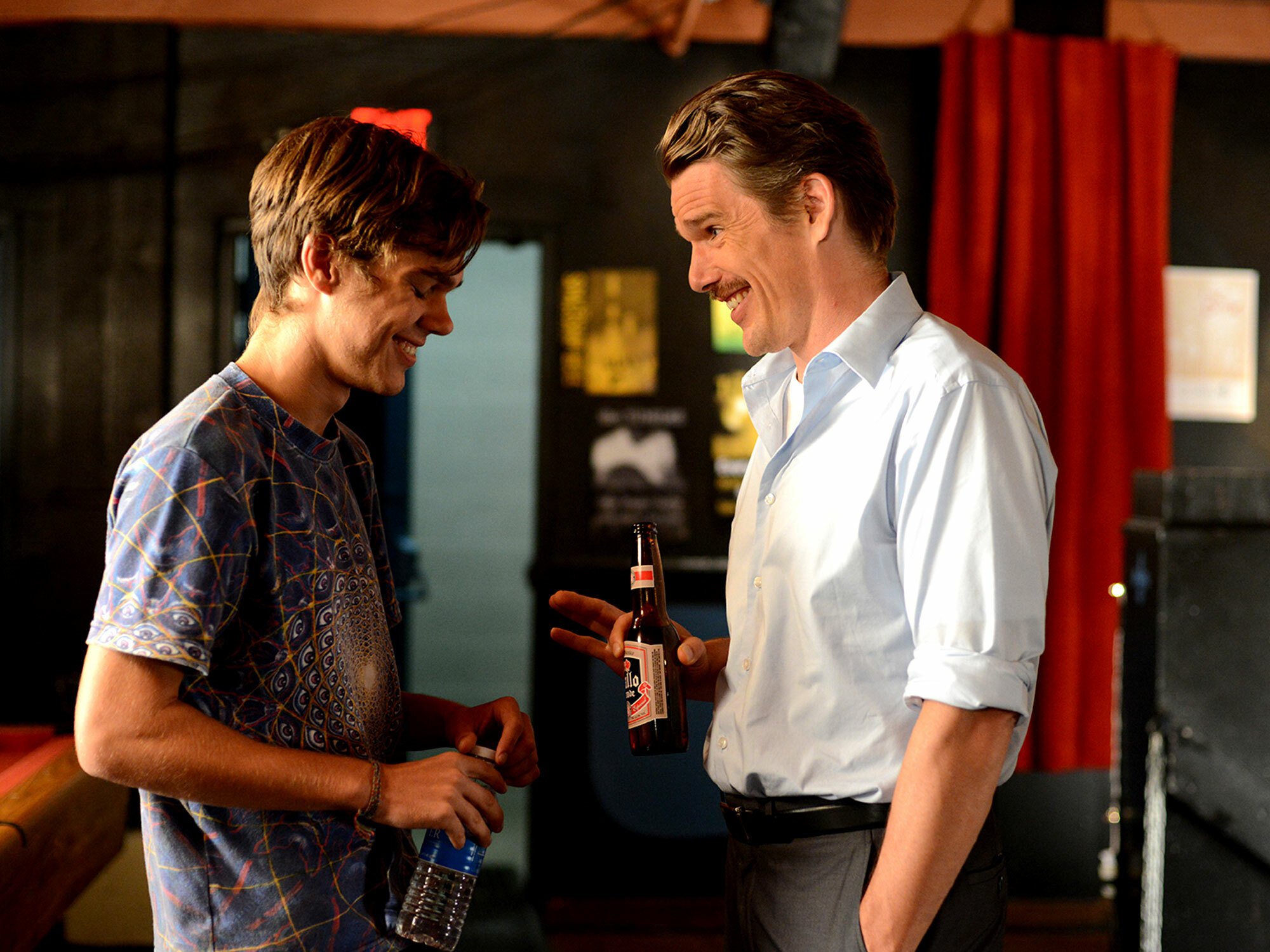 Two men chatting and laughing in a bar, one holding a bottle.
