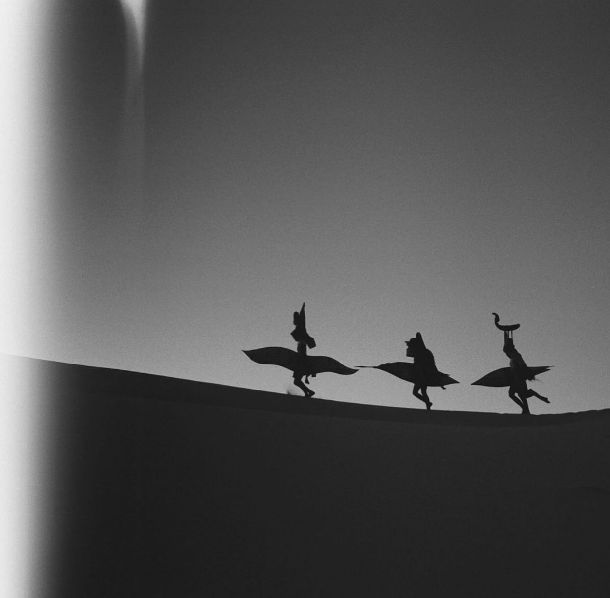 Black and white image showing silhouettes of three people carrying surfboards walking along a ridge against a gradient grey sky.
