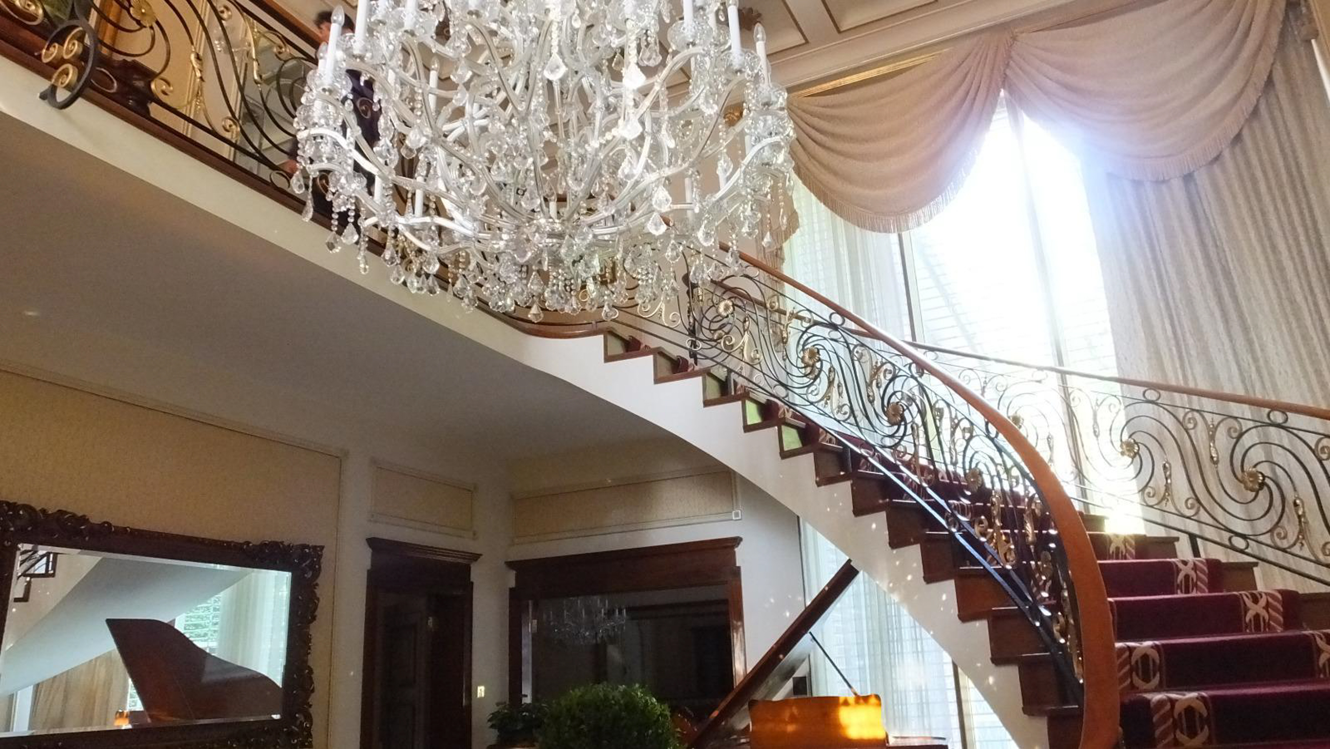Grand staircase with ornate metal railings, crystal chandelier above, cream curtains, marble steps, and modern interior visible below.