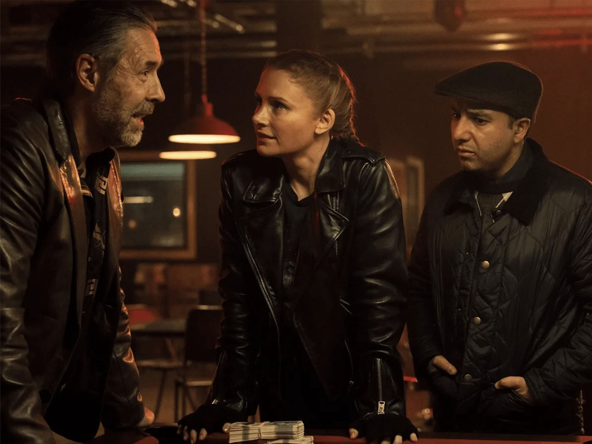 Three adults in dark leather jackets standing in a dimly lit room, one woman and two men