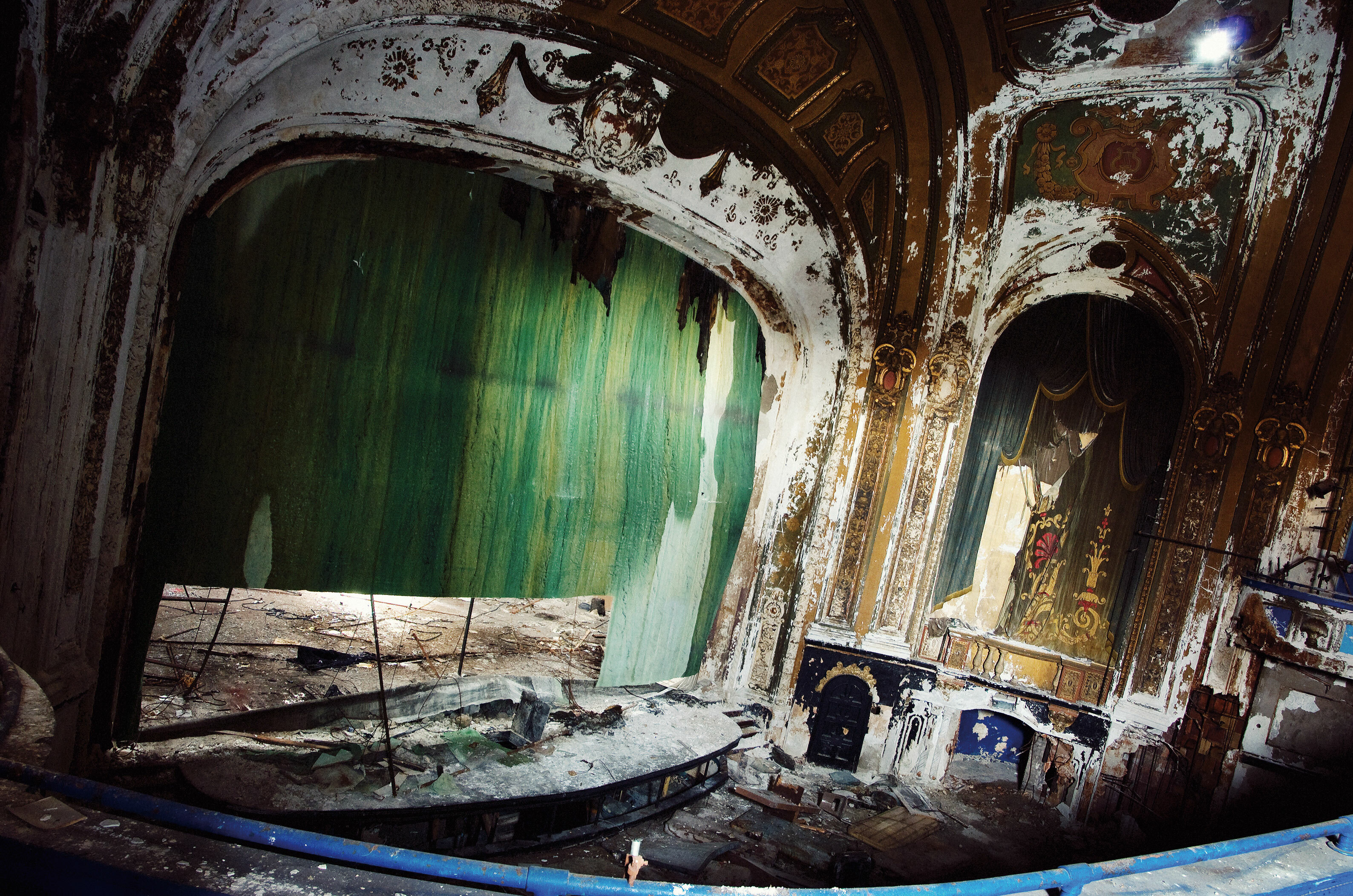 Dilapidated once-grand theatre auditorium with ornate decaying arches, green curtains, and debris on floor.