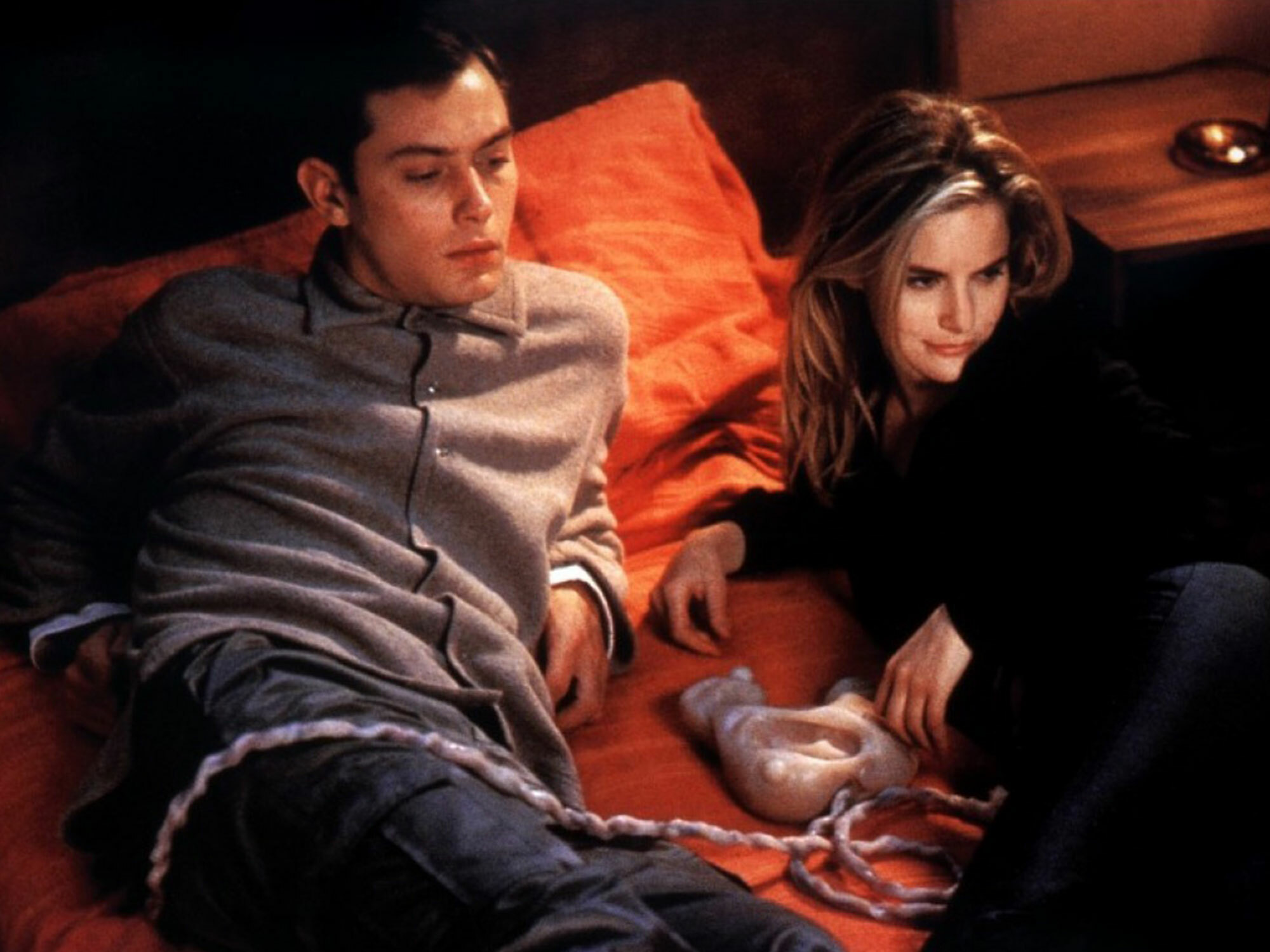 Two individuals, a man and a woman, sitting on a couch in a dimly lit room. The man appears to be in a relaxed, contemplative pose, while the woman is leaning towards him.