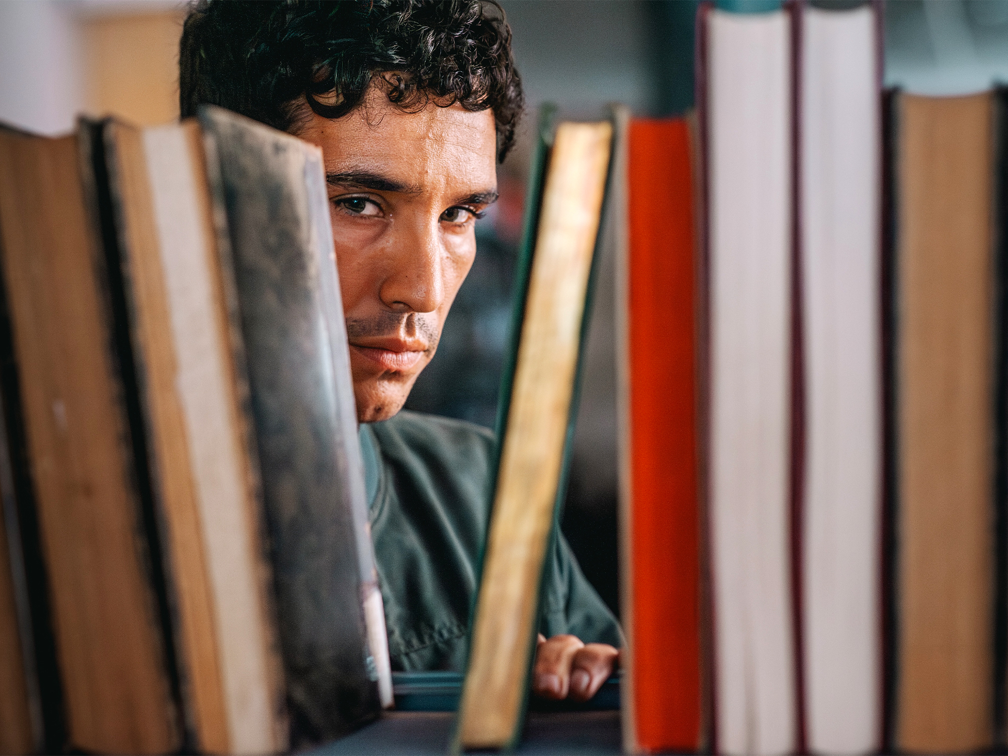 Man with dark curly hair peering through gap between books on shelf, wearing green shirt, books in various colours surrounding him.