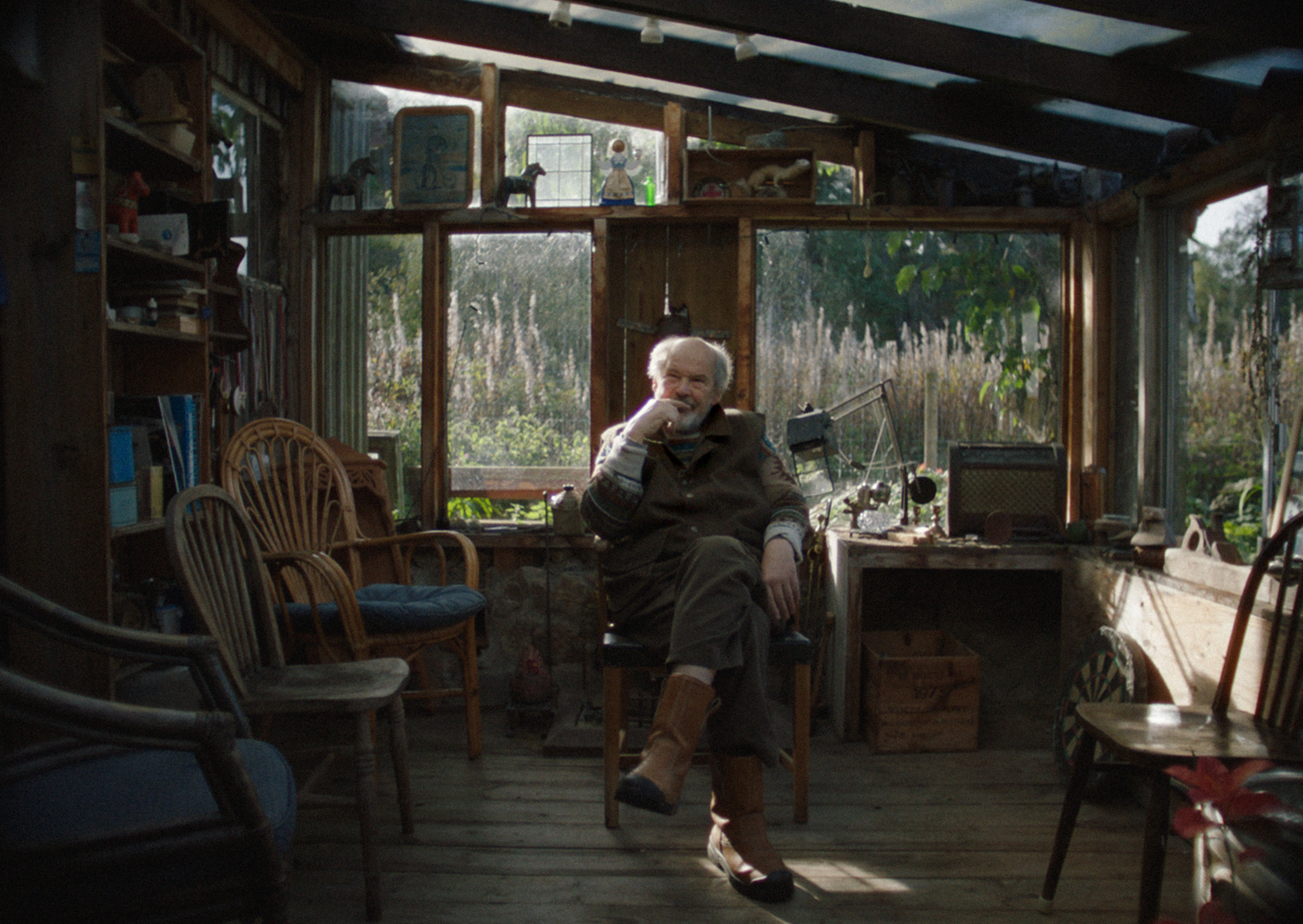 Elderly man with white beard sits in cluttered glass-walled room with wooden furniture, papers, and natural light streaming through windows.