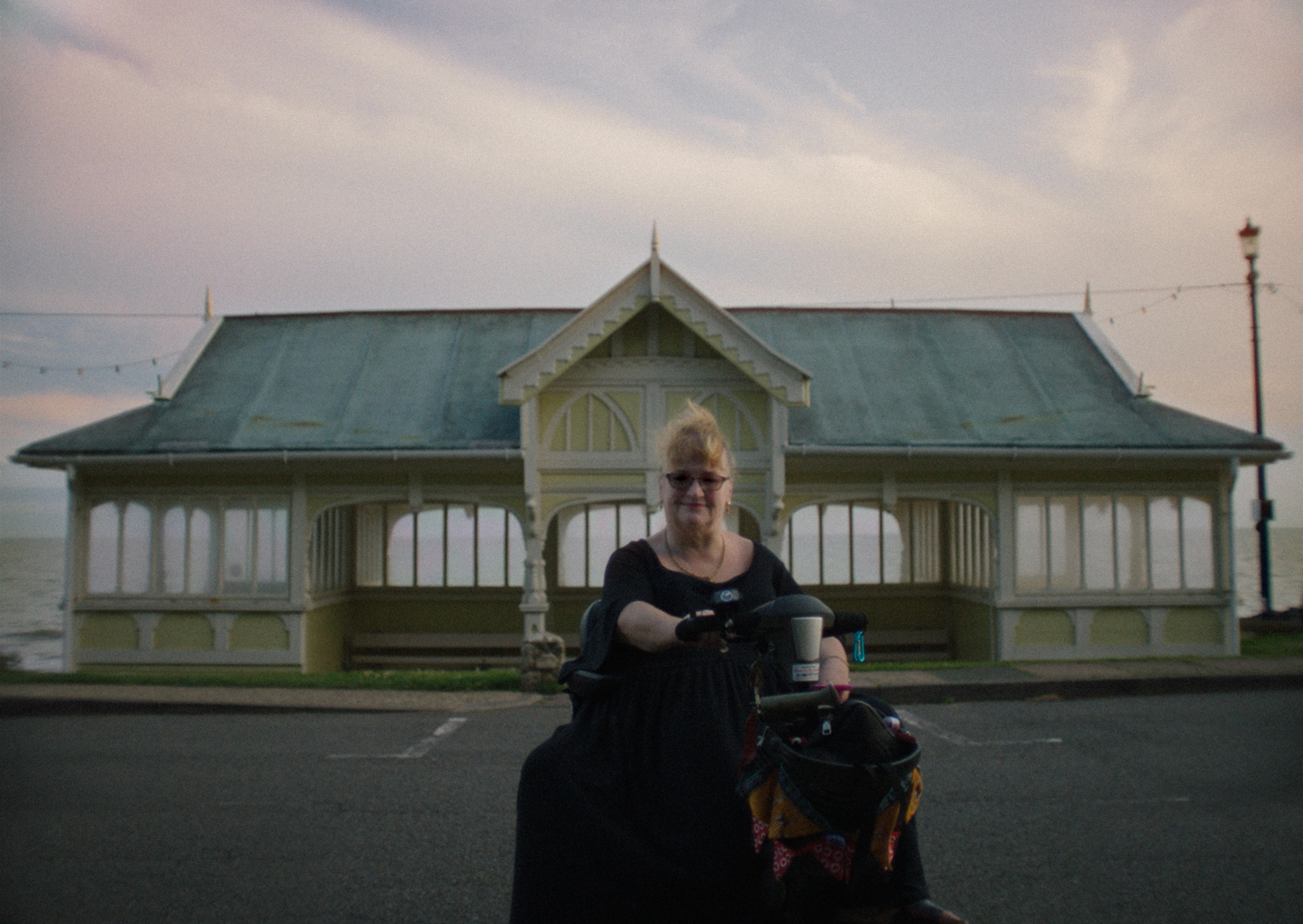 Person in wheelchair in front of Victorian-style pavilion with green roof, white walls, and ornate details under cloudy sky.