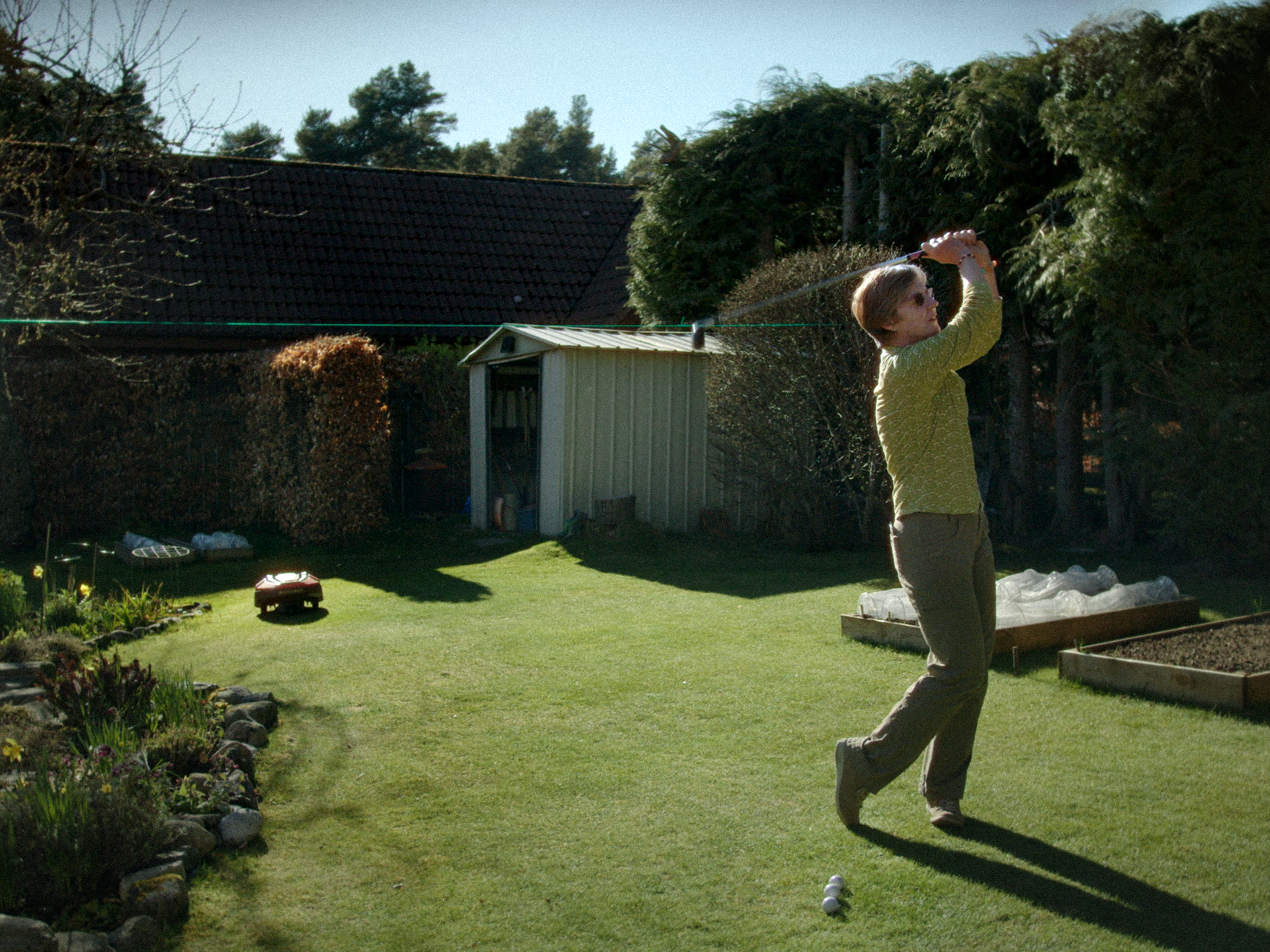 Man in yellow shirt mid-golf swing on garden lawn with grey shed, stone walls, and trees in background.
