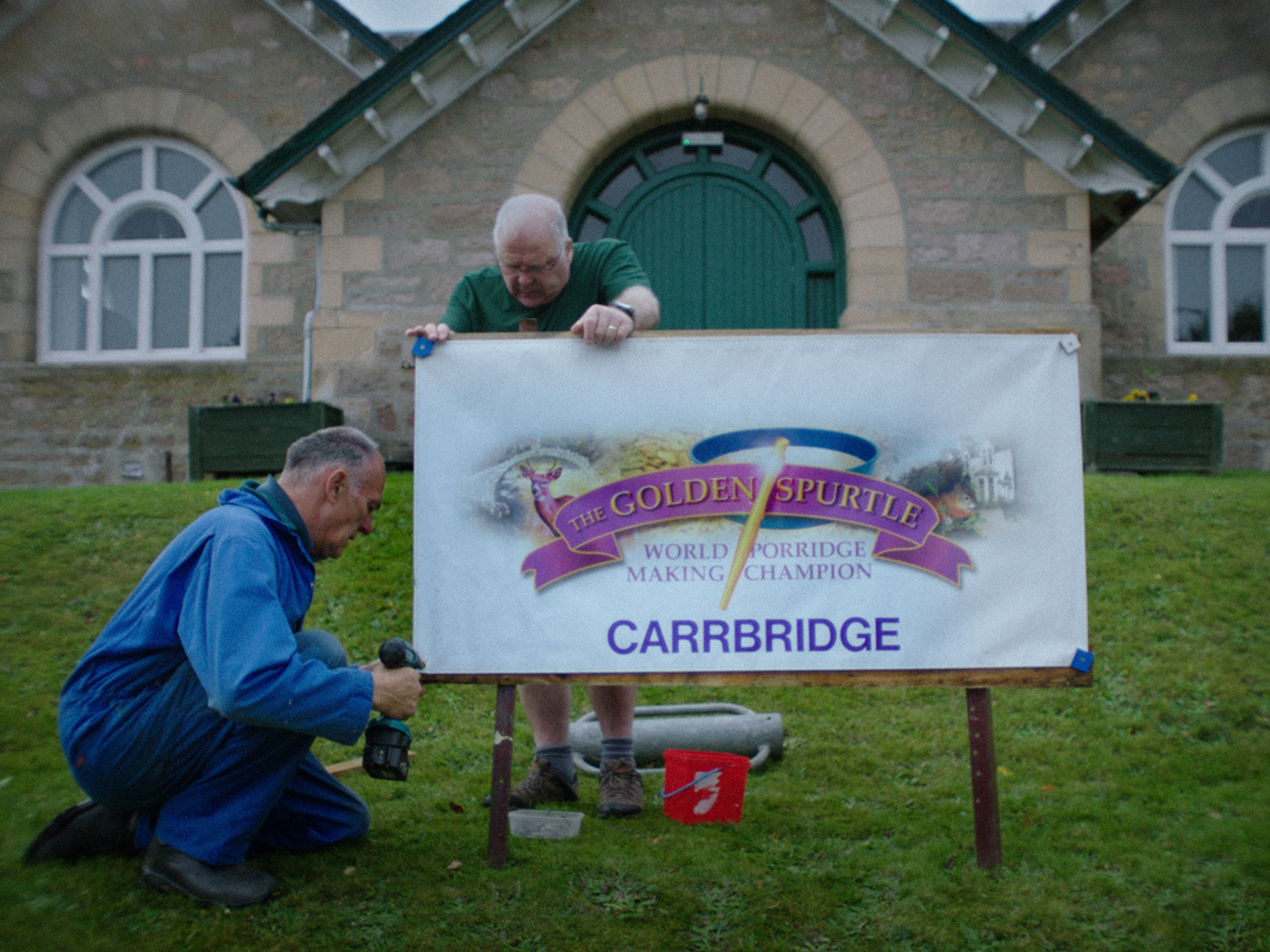 Two men installing white sign reading "The Golden Spurtle World Porridge Making Championship Carrbridge" in front of stone building with green door.