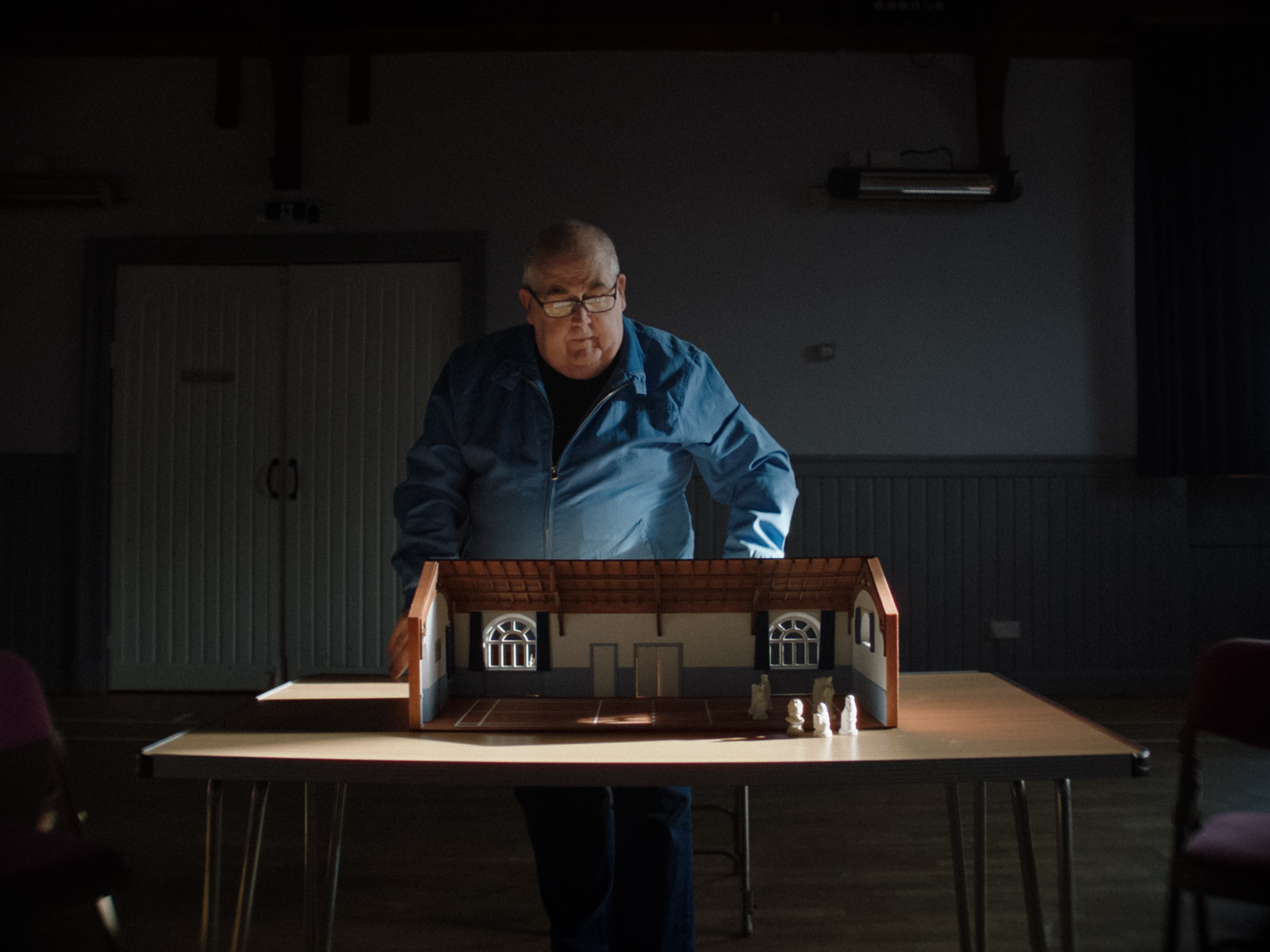 Elderly man in blue jacket examining detailed dollhouse on table in dimly lit room with window light illuminating the scene.