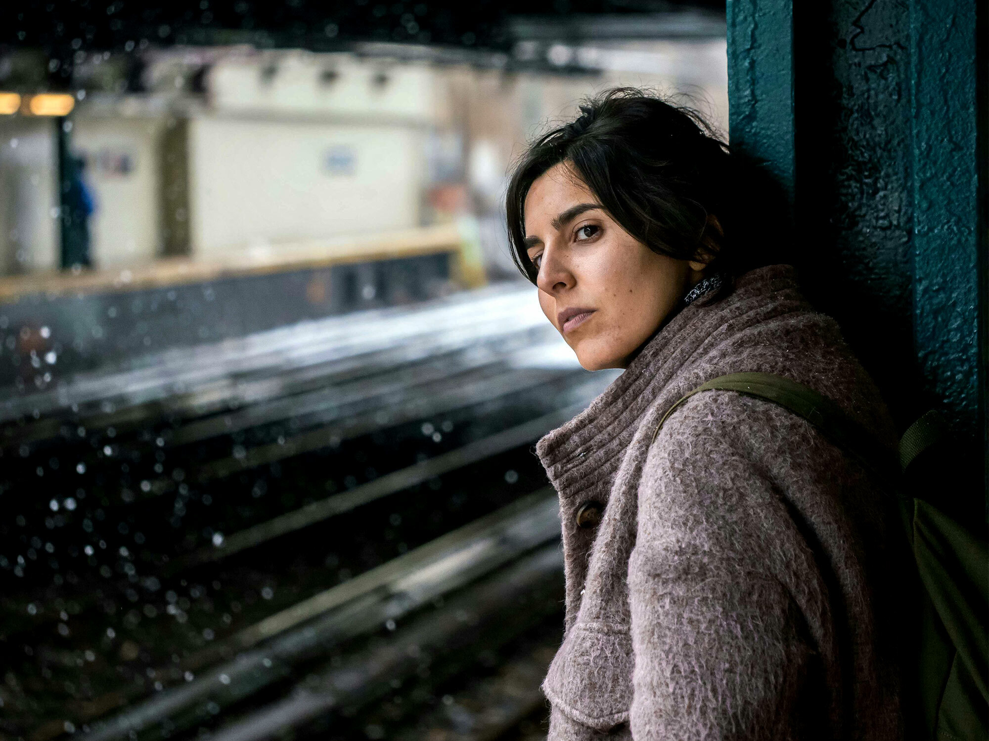 A woman with dark hair wearing a knitted brown jumper stands against a wall, looking pensive and gazing into the distance.