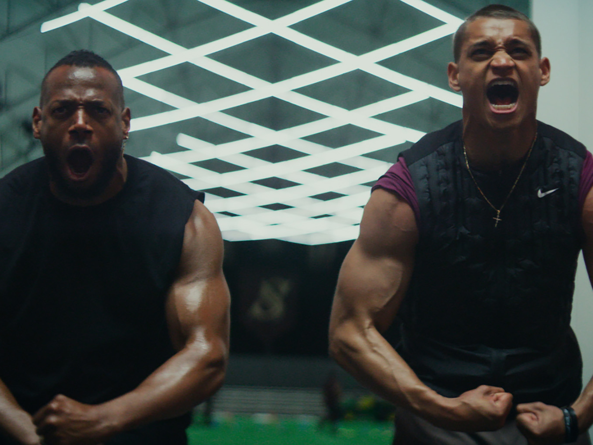 Two men in dark athletic shirts against green and white geometric patterned ceiling, one shouting with mouth open.