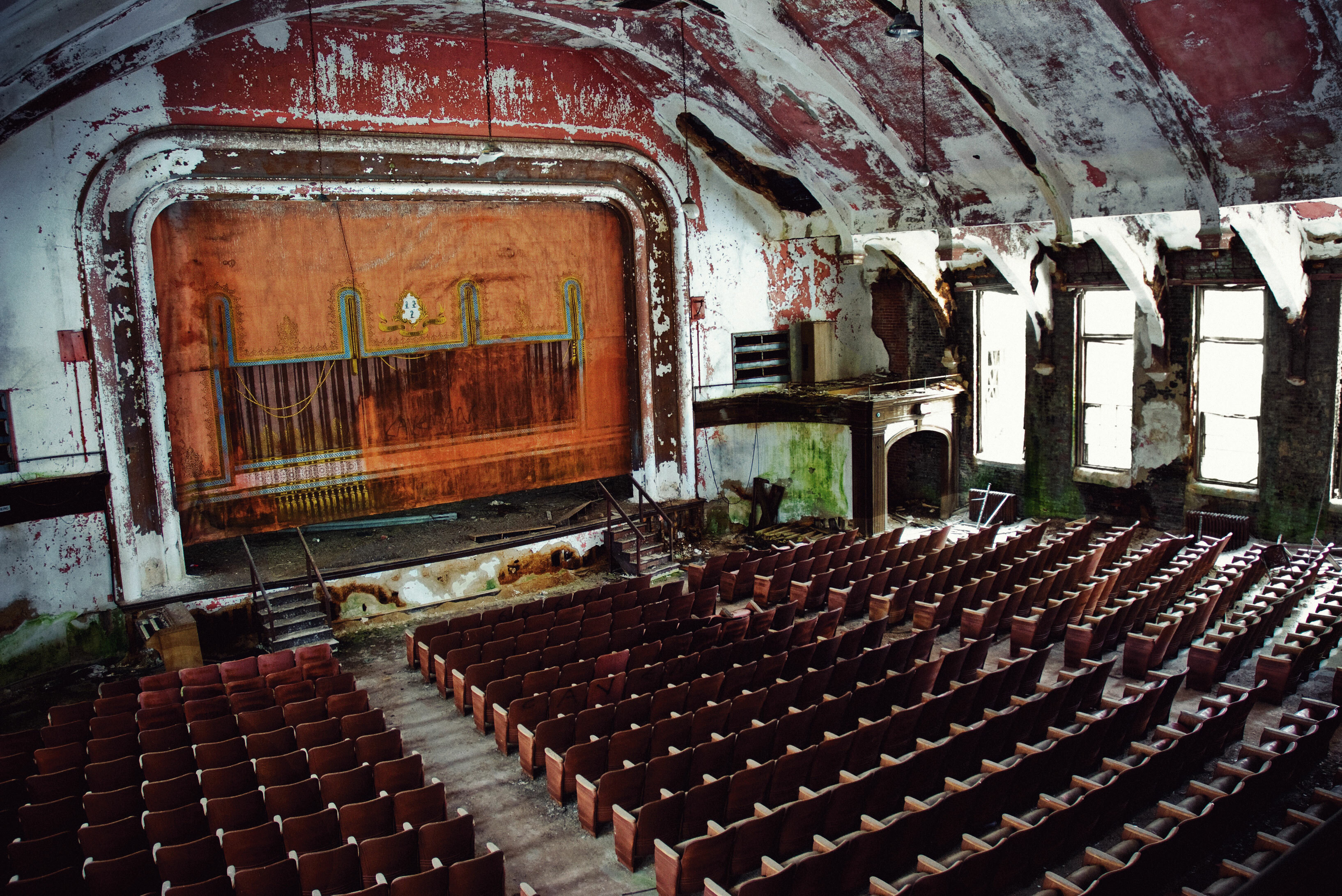 Dilapidated interior of an abandoned theatre with rows of seats and a large stage at the far end.