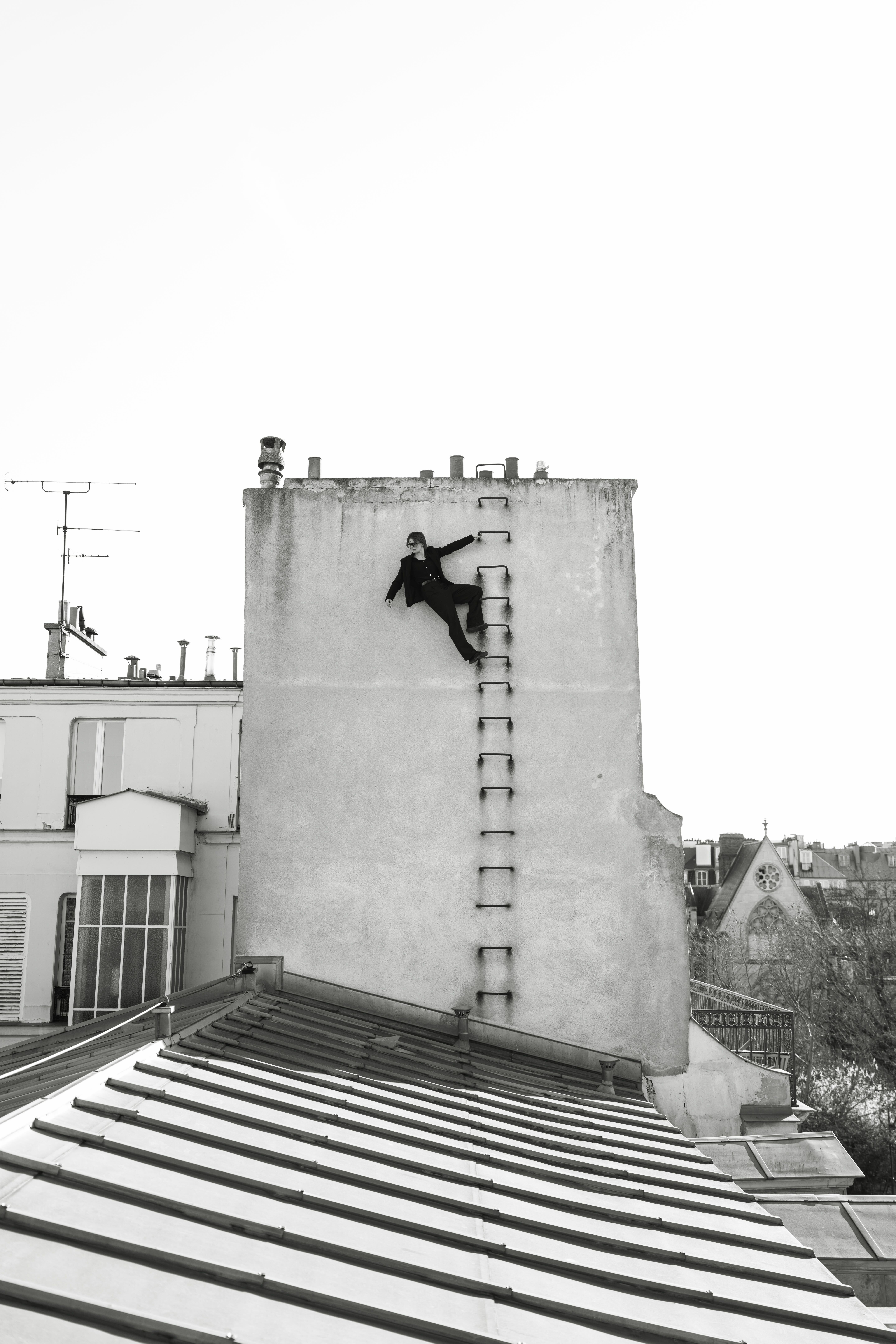 Street art mural of black silhouette figure climbing ladder on white building wall, viewed from tiled rooftop in black and white.