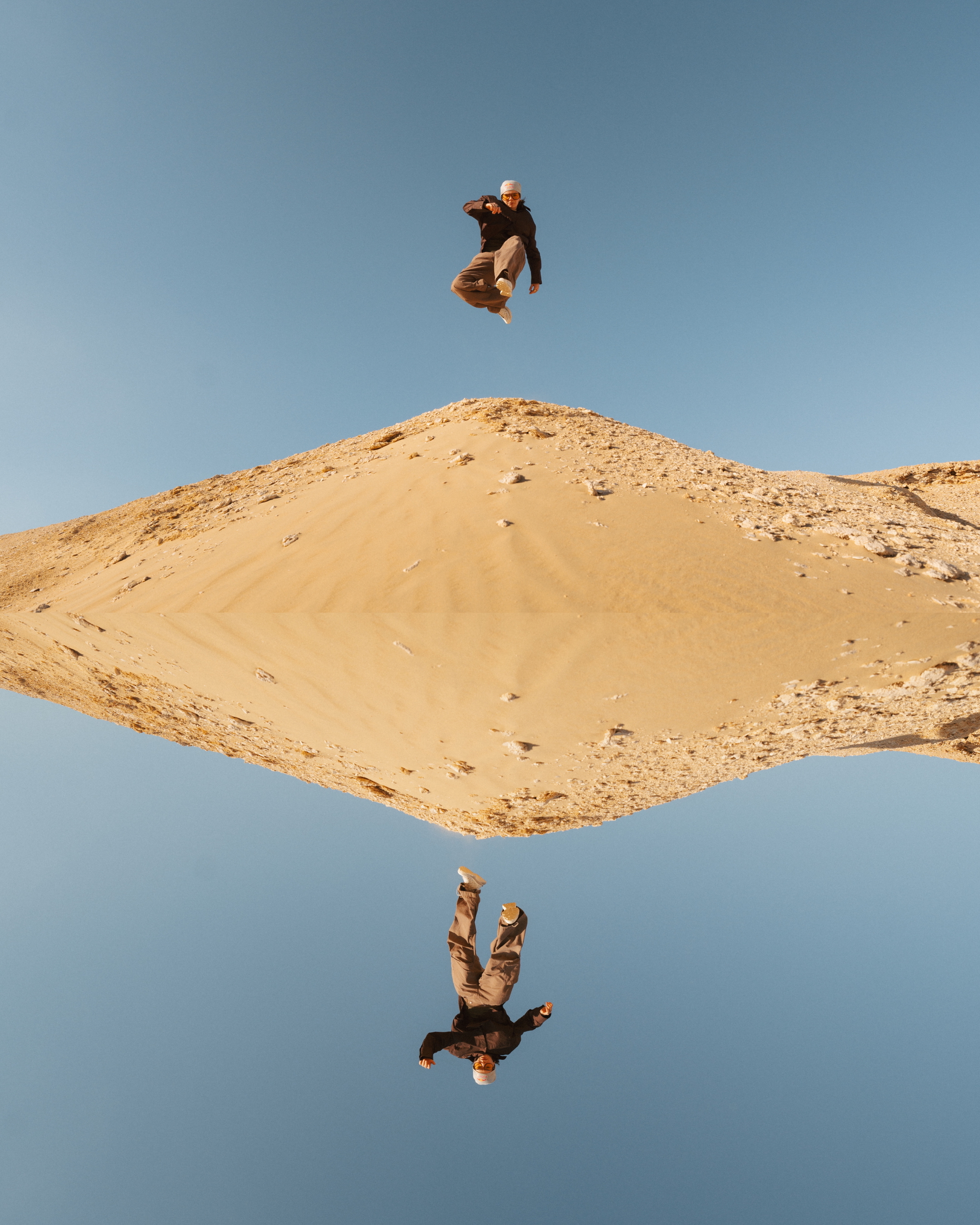 Person jumping above sandy hill with clear blue sky, their reflection visible in still water below creating mirror effect.