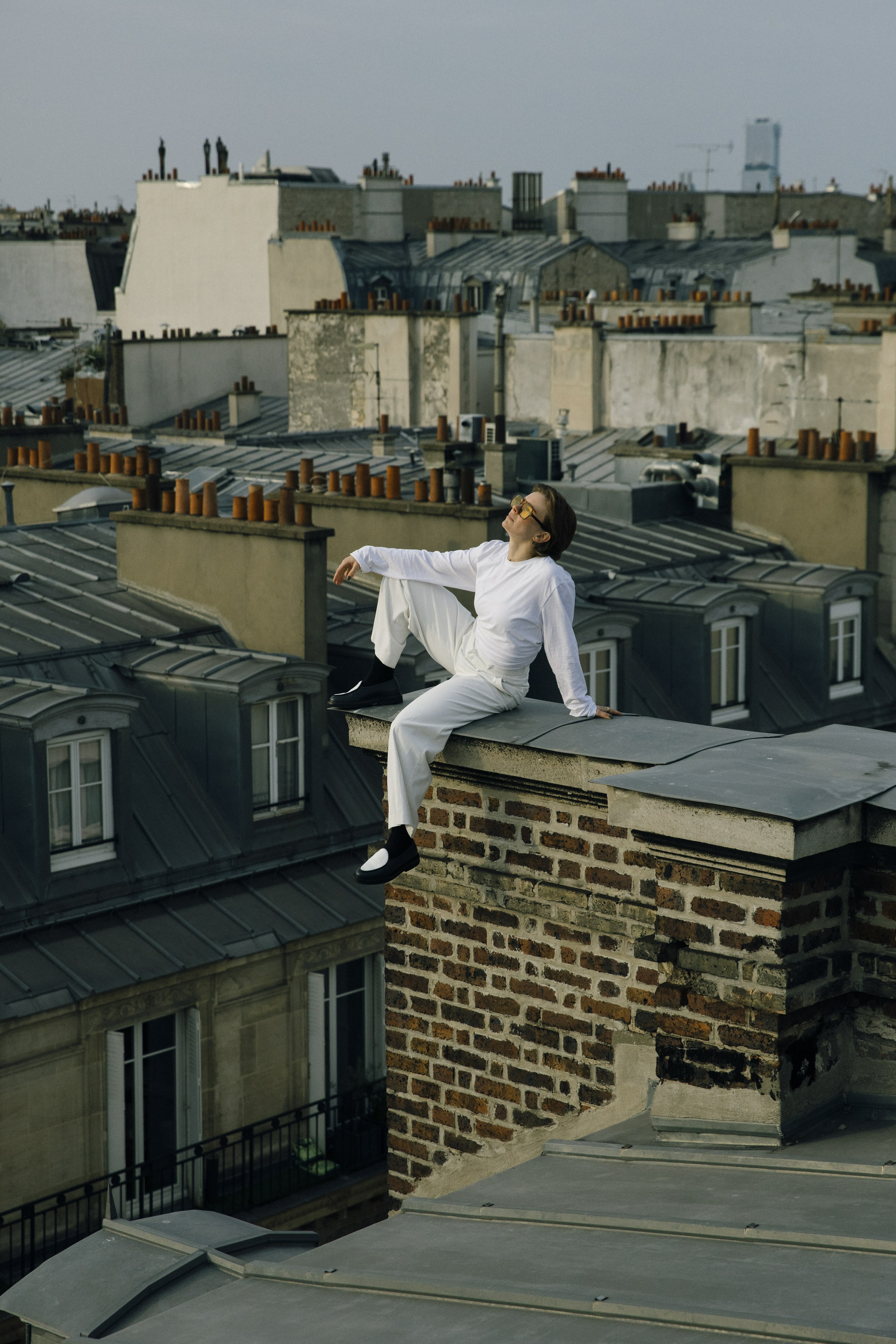 Person in white clothing sits on brick chimney overlooking Parisian rooftops with grey slate tiles and cream-coloured buildings.