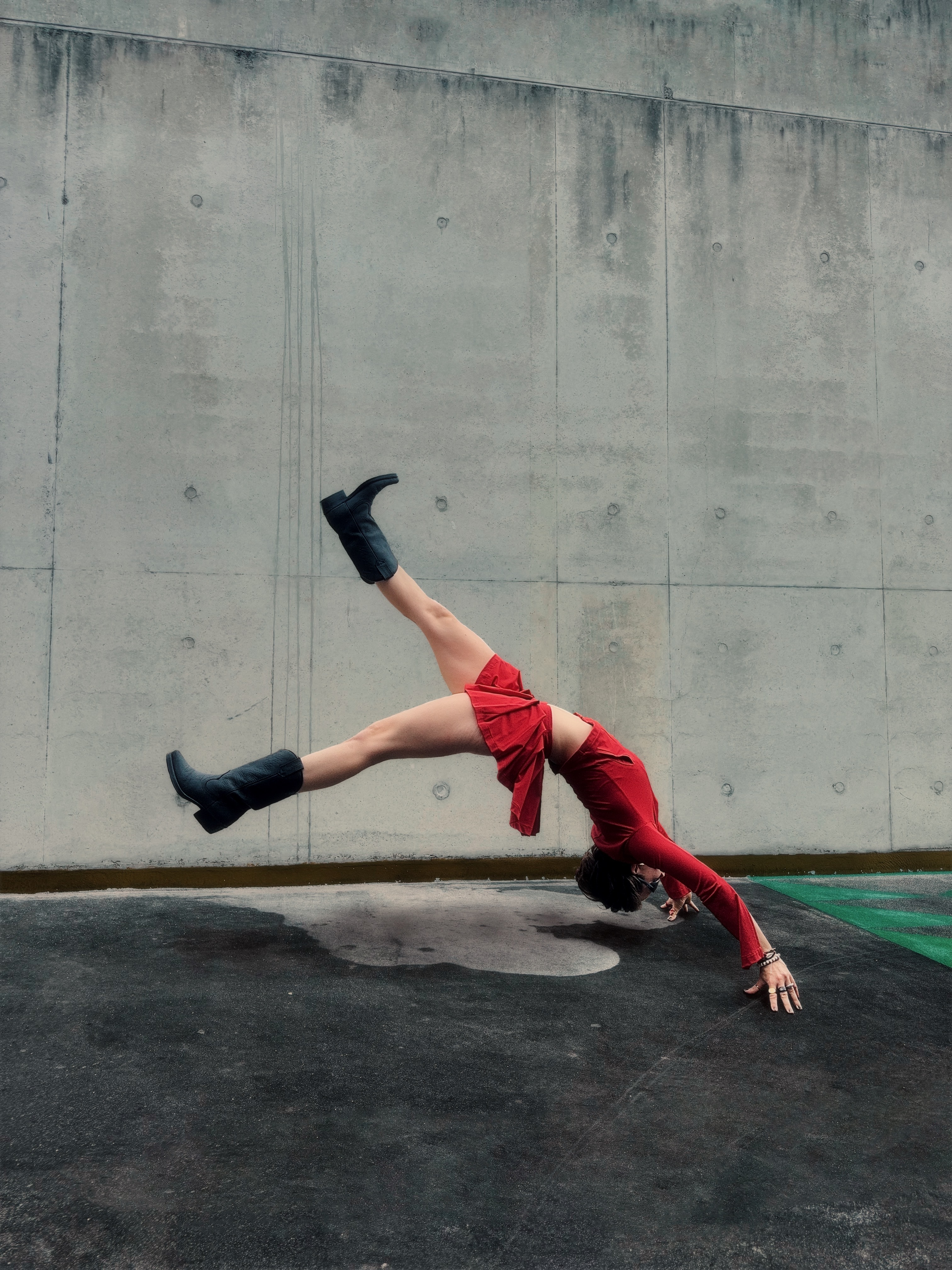Person in red outfit performing headstand with legs extended, black boots, against weathered concrete wall background.