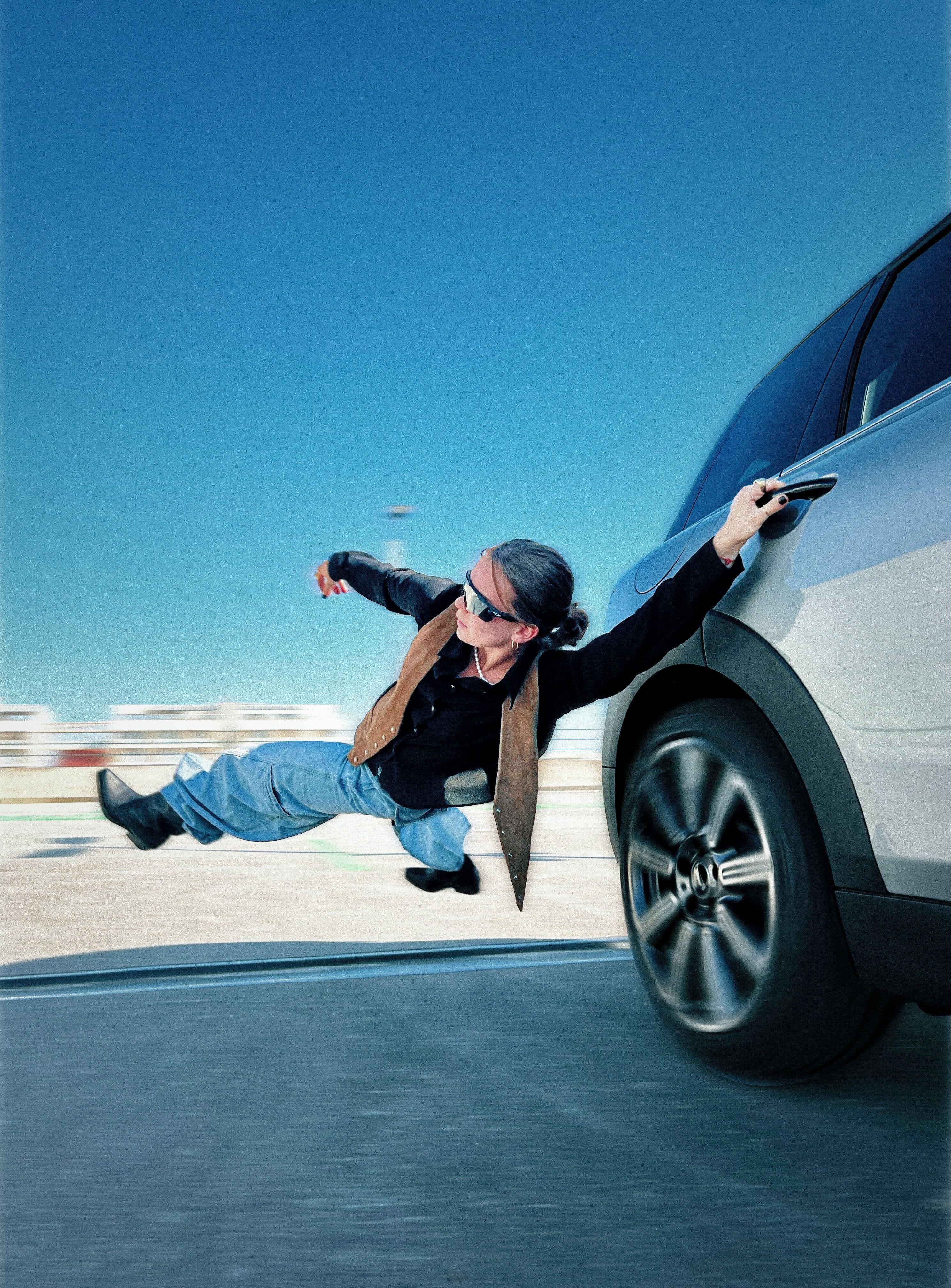 Woman in sunglasses leaning horizontally out of moving blue car door, legs extended, against clear blue sky and blurred road surface.