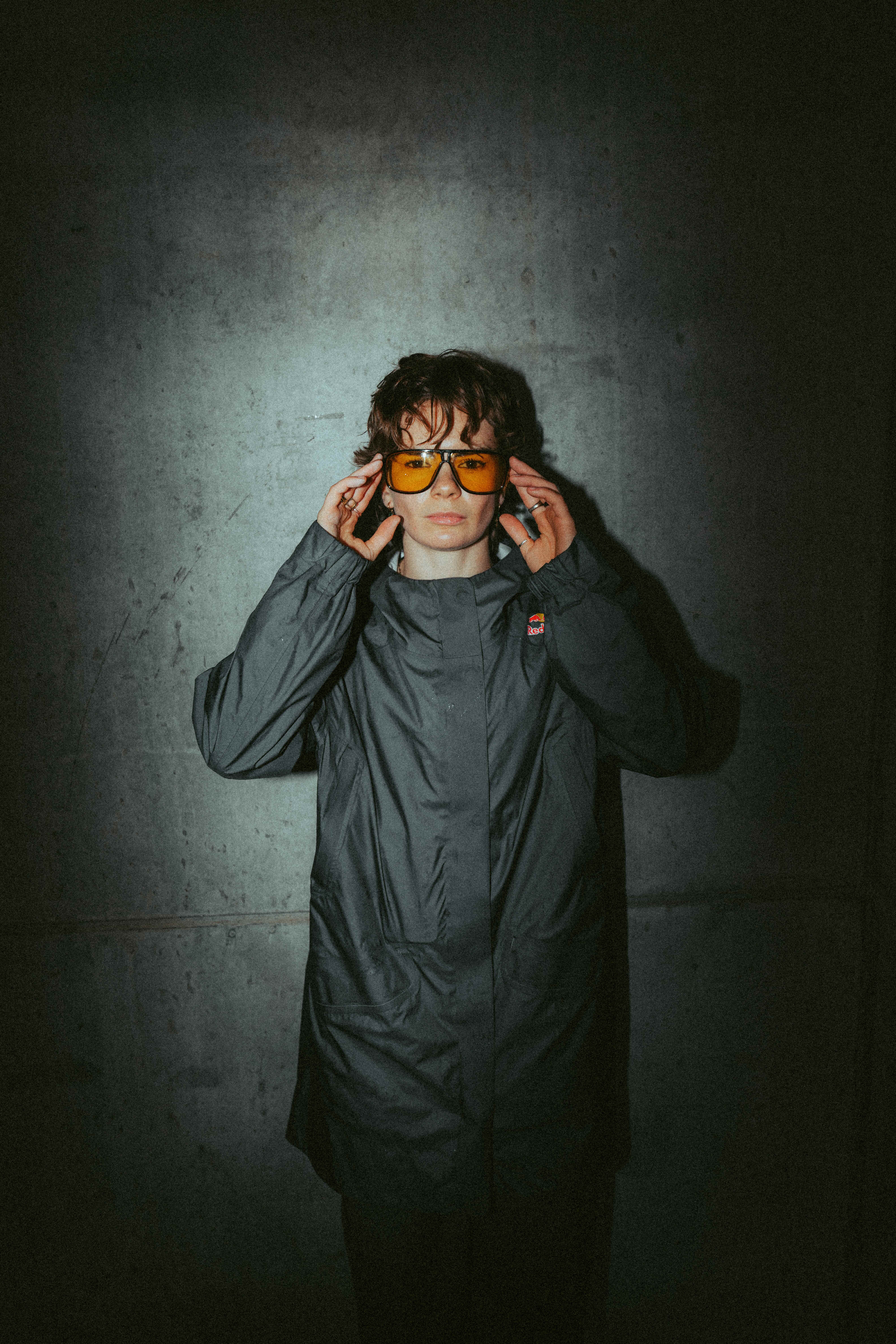 Person with tousled hair wearing orange-tinted sunglasses and dark jacket, hands raised to temples, against concrete wall background.