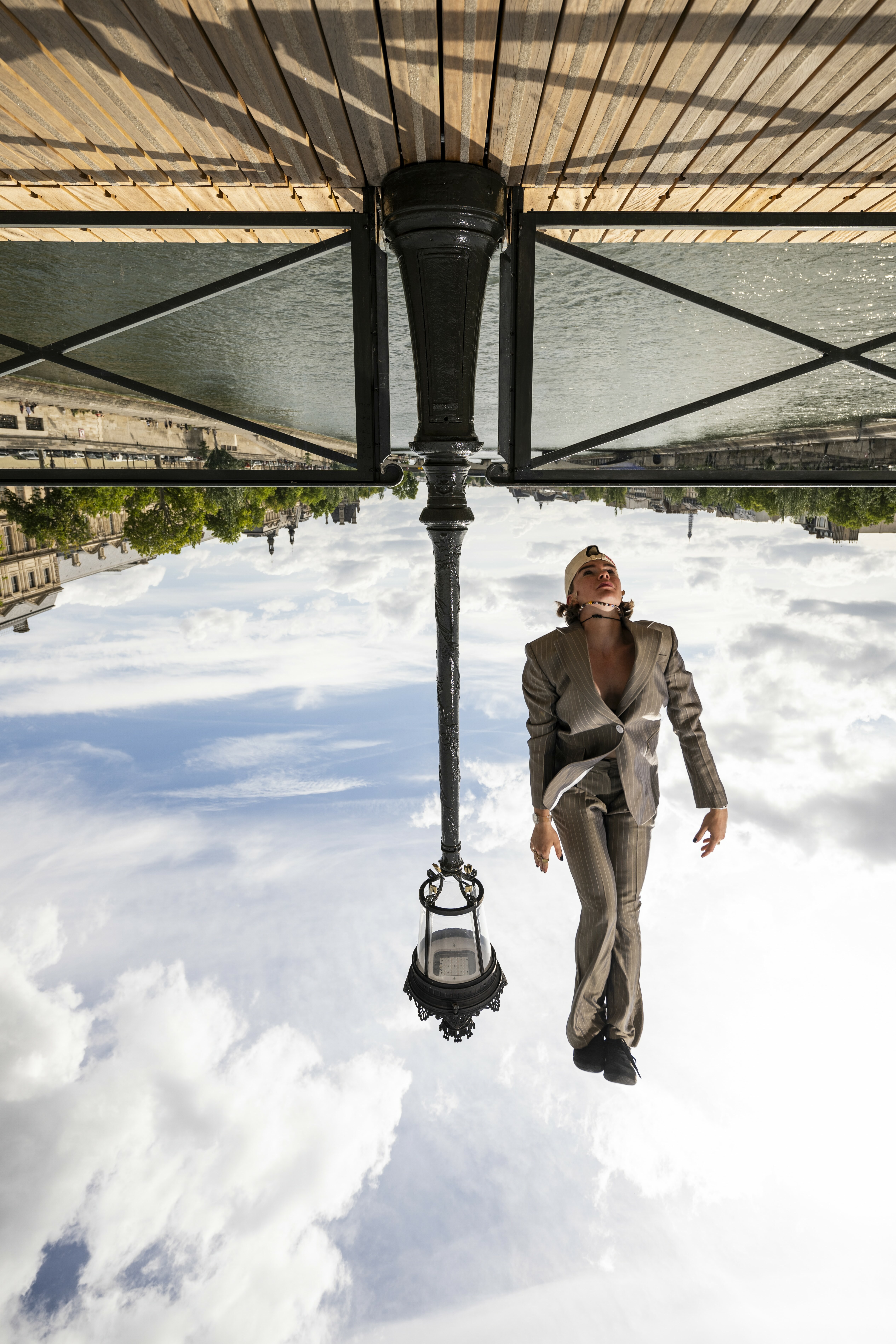 Man in brown suit walks upside down on mirrored ceiling beneath ornate lamp post, with cloudy sky and architectural details reflected.