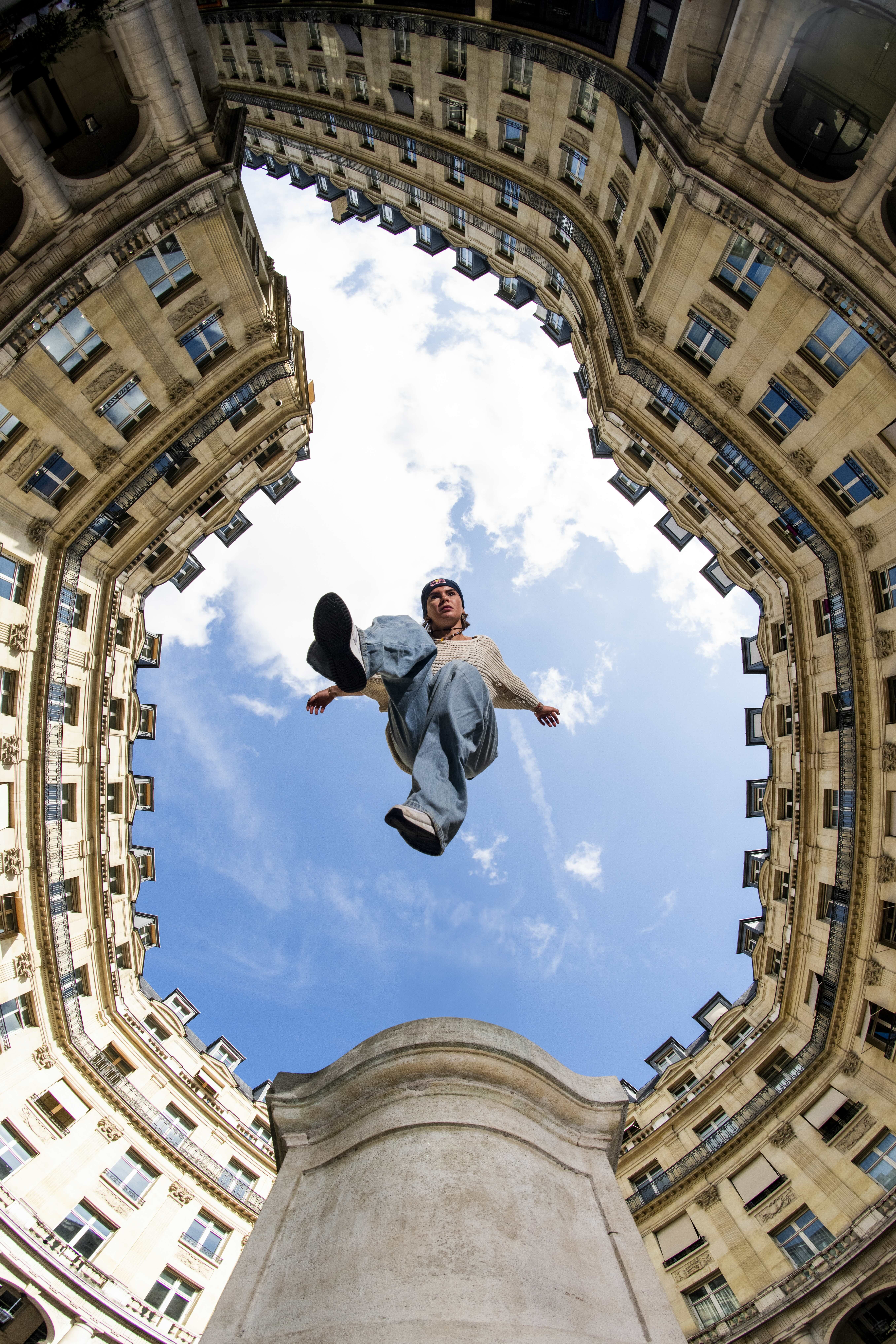 Person jumping mid-air in courtyard surrounded by cream stone buildings with blue windows, shot from below against cloudy sky.