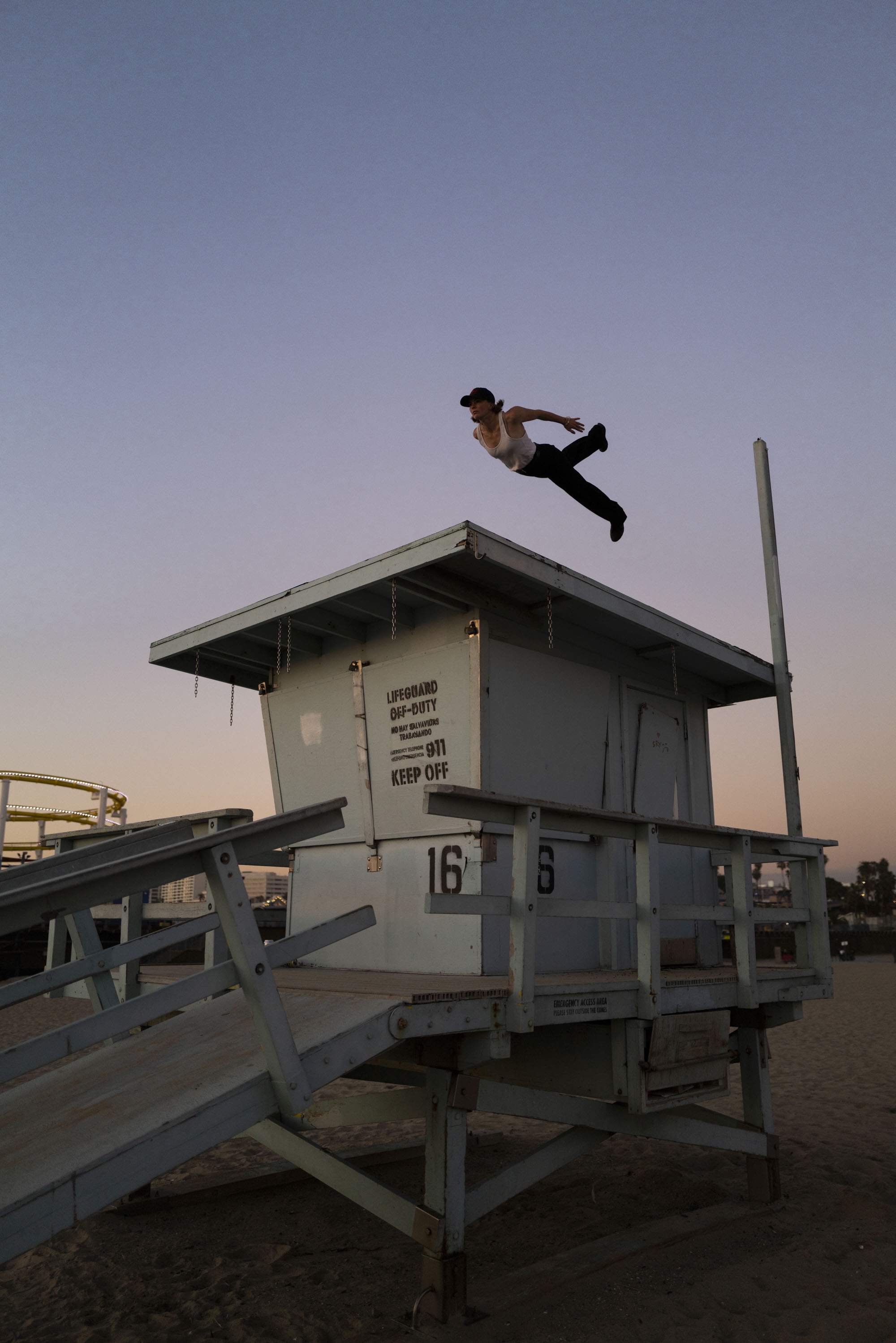 Person jumping off white lifeguard tower numbered 16 against purple-pink sunset sky on beach.