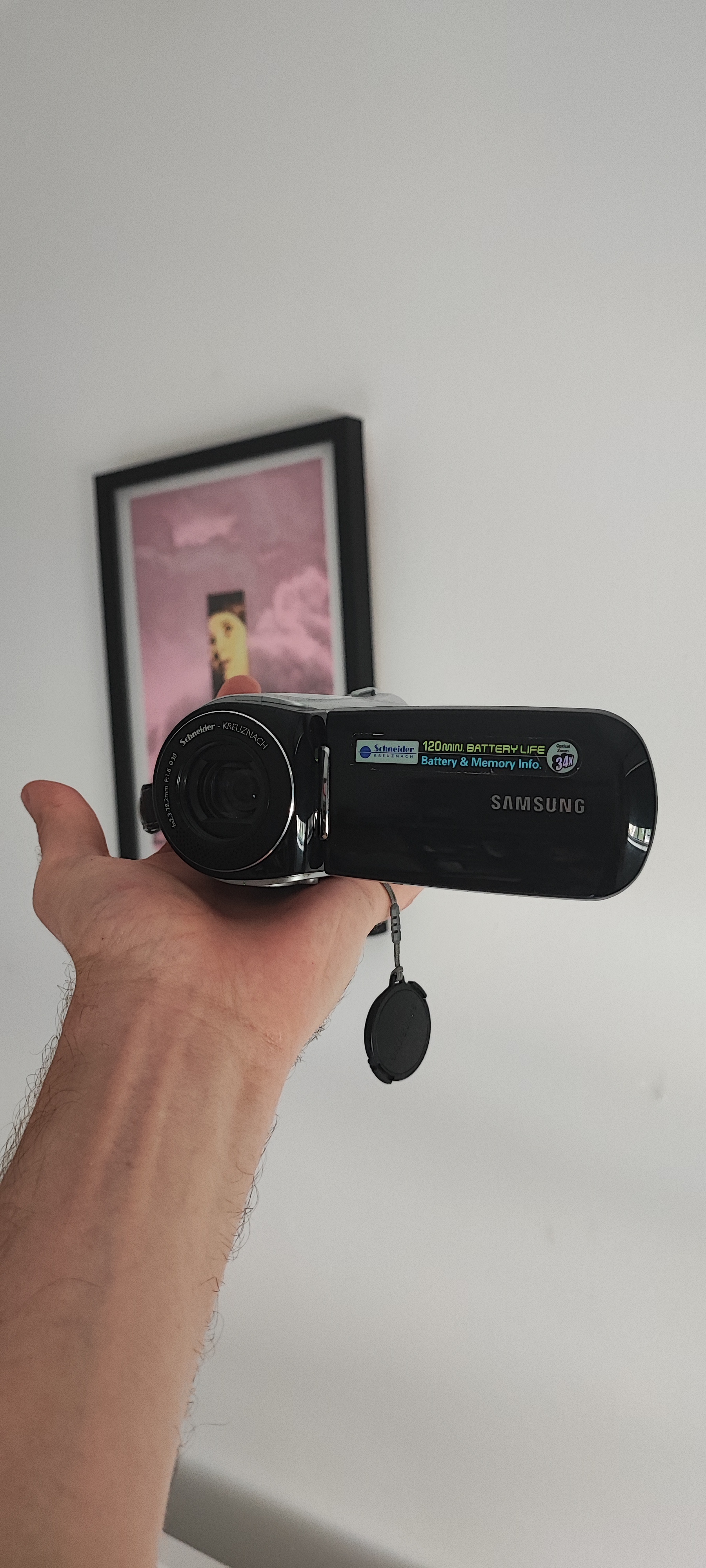 Hand holding black Samsung camcorder with lens cap dangling, framed artwork with pink background visible on white wall behind.