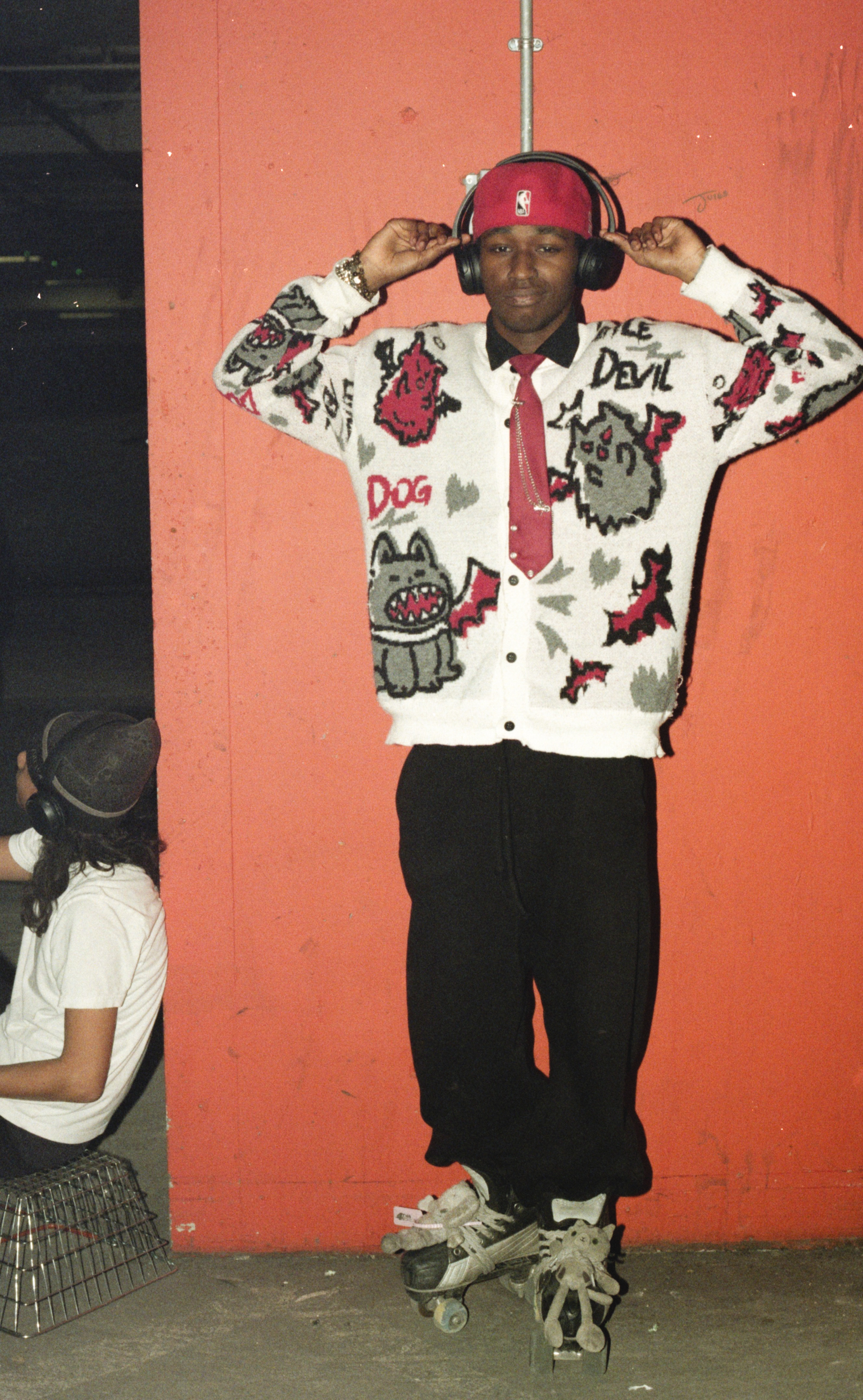 Man wearing headphones, red cap, white shirt with cartoon dog graphics, and black trousers against orange wall.
