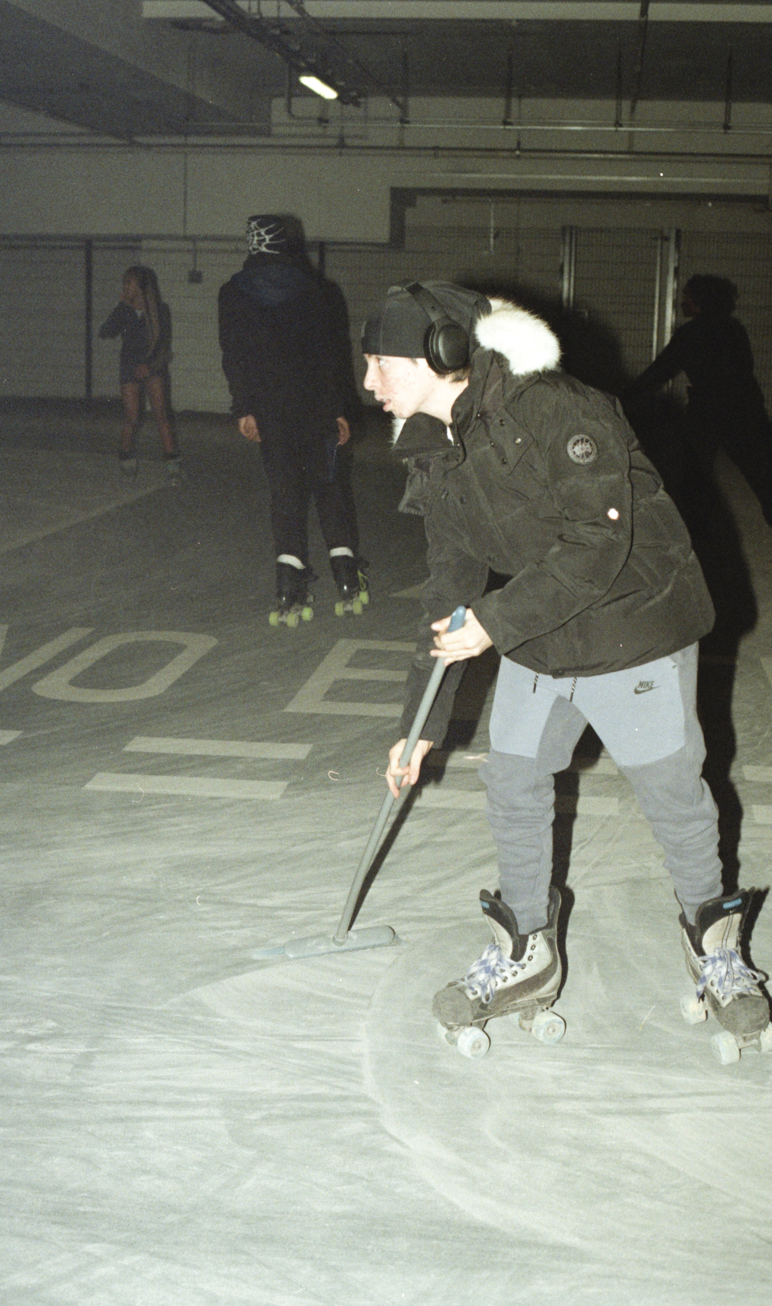 Person in green jacket and beanie playing roller hockey in indoor rink, holding stick whilst on roller skates with others nearby.