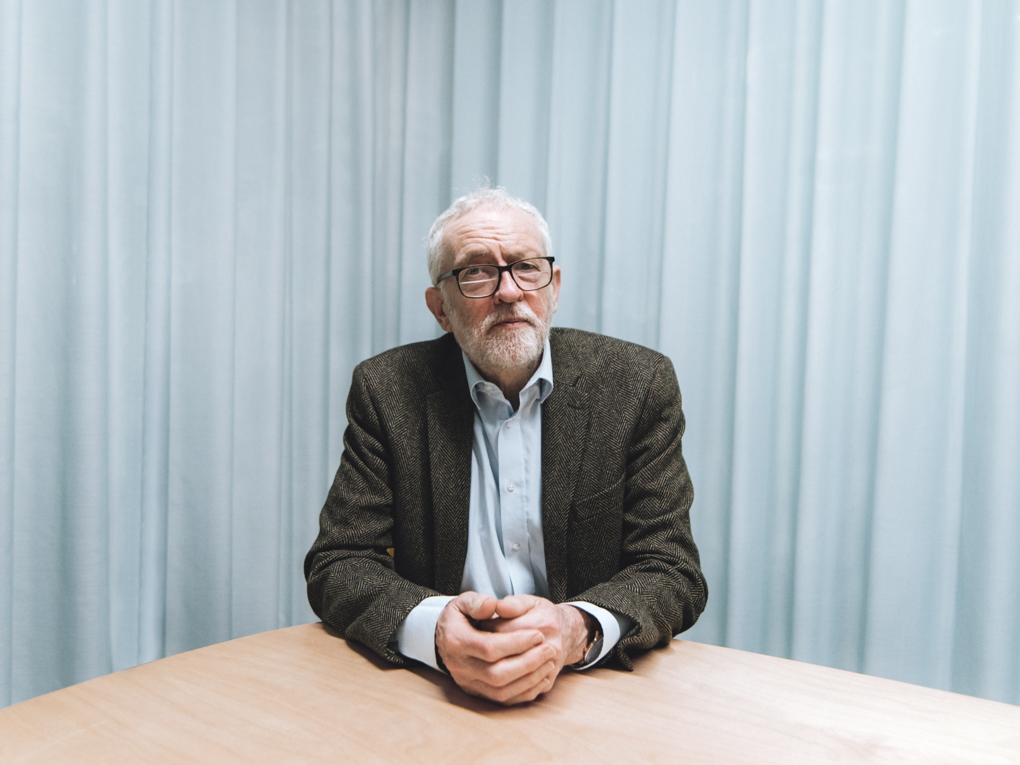 Elderly man with white hair and beard wearing glasses, dark jacket and light shirt, sitting at wooden table against white curtains.