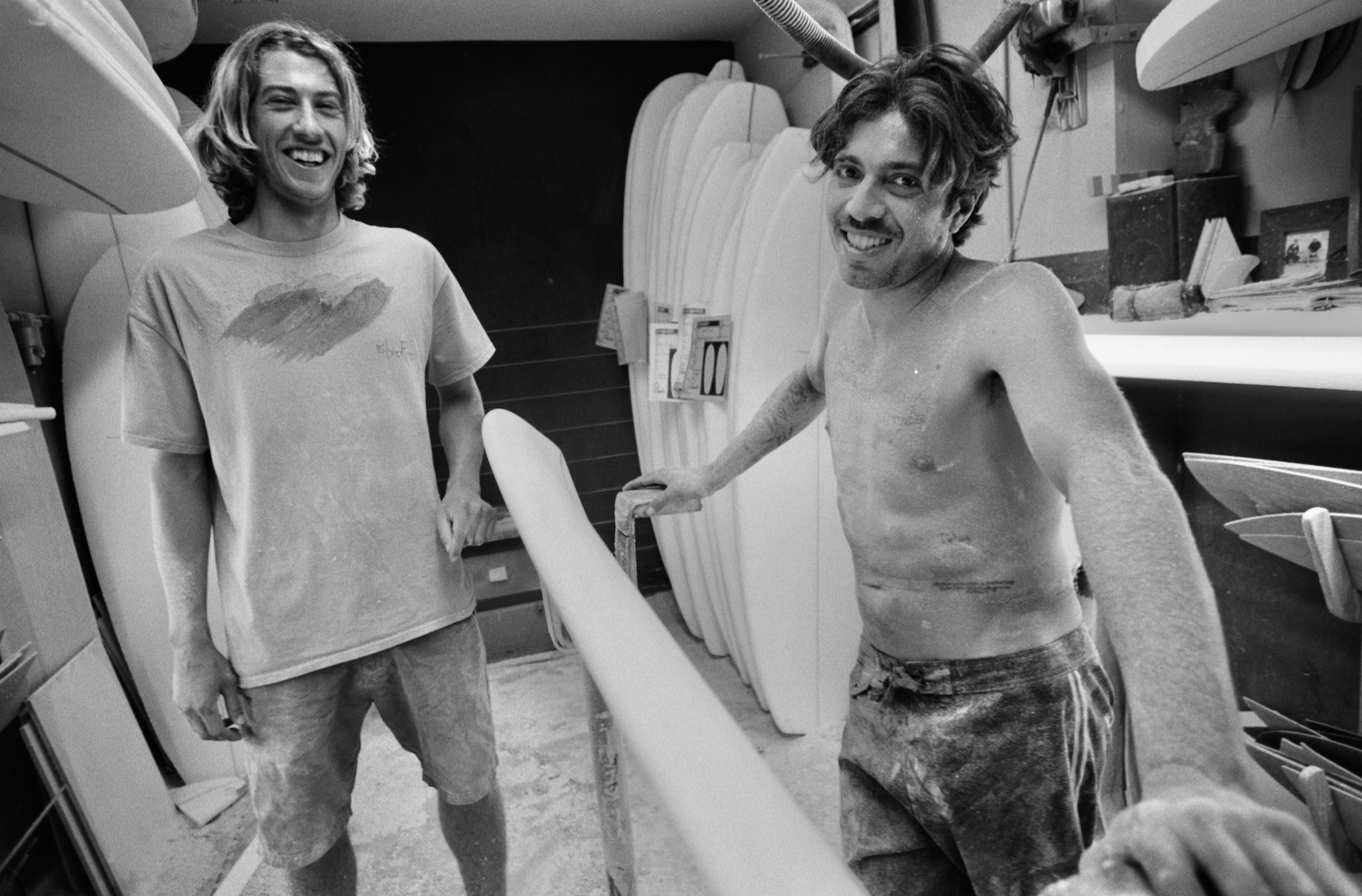 Two men in surfboard workshop, one shirtless with messy hair, other in t-shirt, standing amongst white surfboards and equipment.