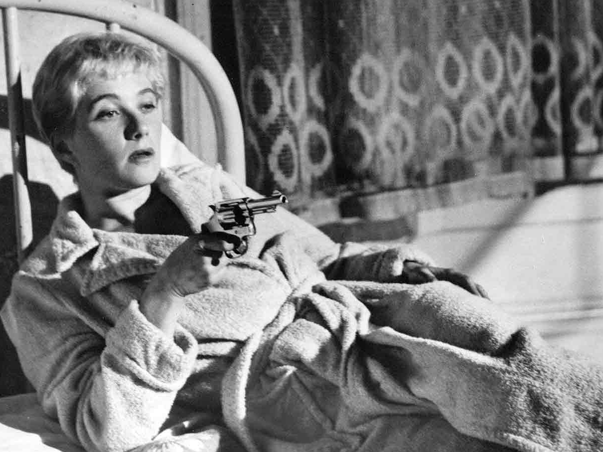 A woman in a robe holding a handgun while lying on a sofa against a patterned wall.