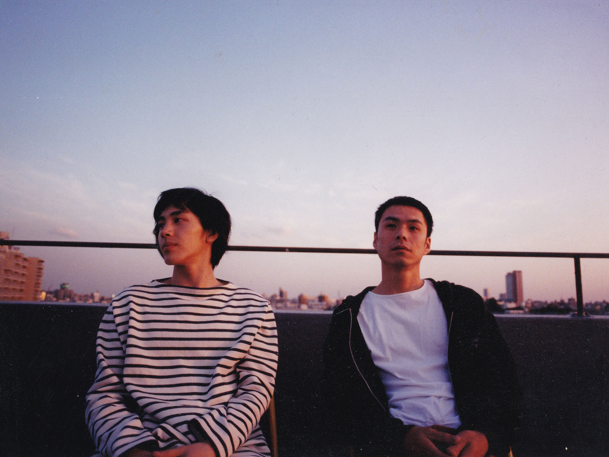 Two young men, one in a striped long sleeve t-shirt and the other in a white t-shirt and black hoodie, sitting on a balcony with the Tokyo cityscape in the background.
