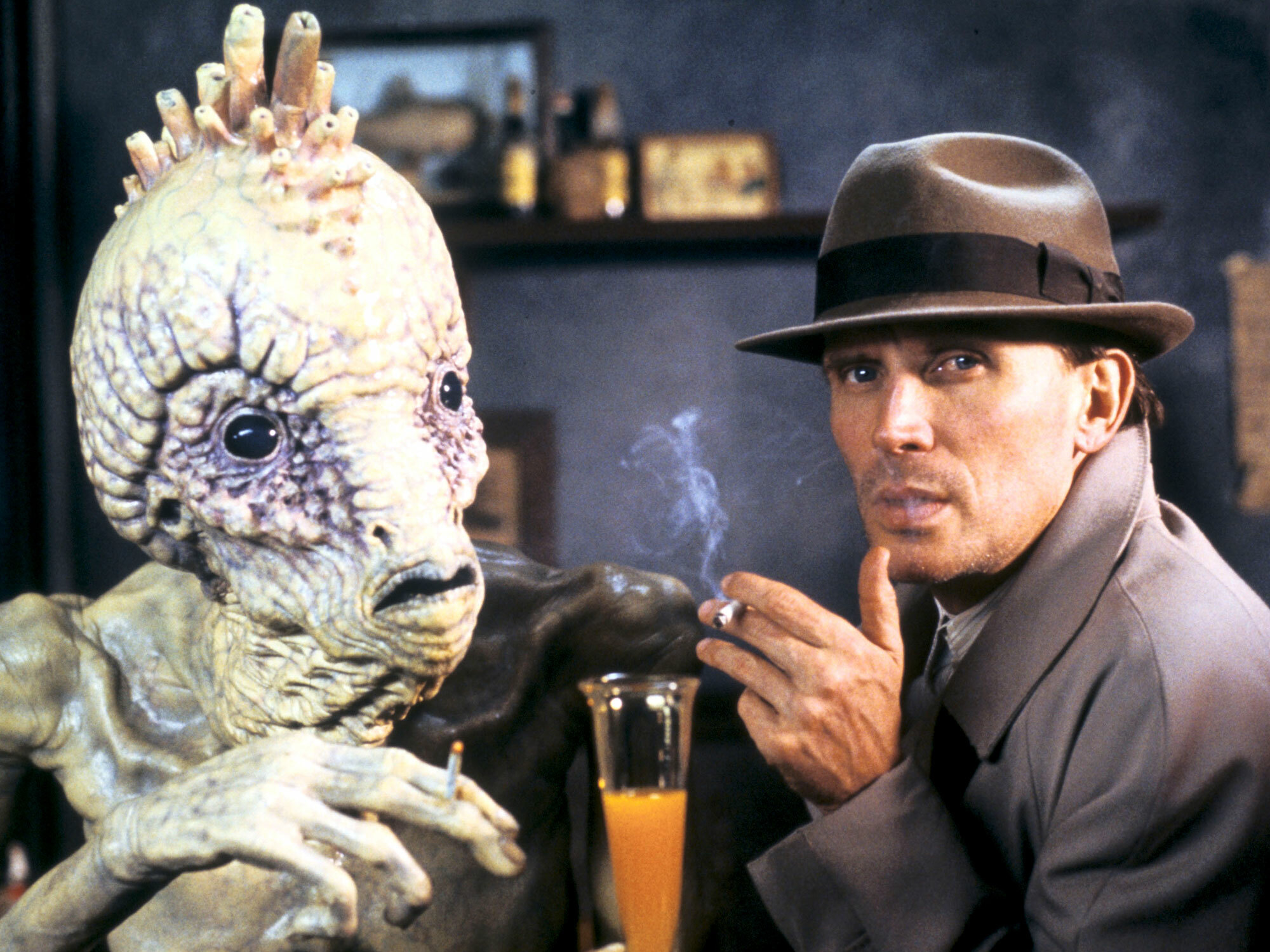 Alien creature conversing with man wearing hat and coat, smoking cigarette.