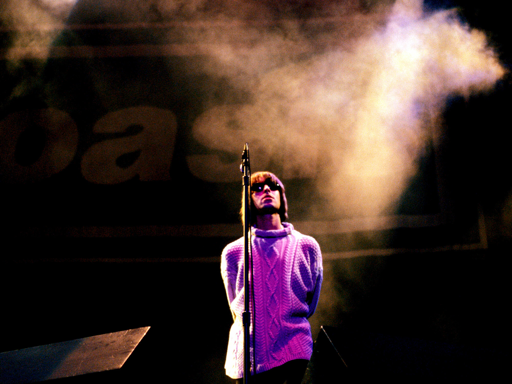 Performer on stage surrounded by smoke, wearing a purple jumper and sunglasses.