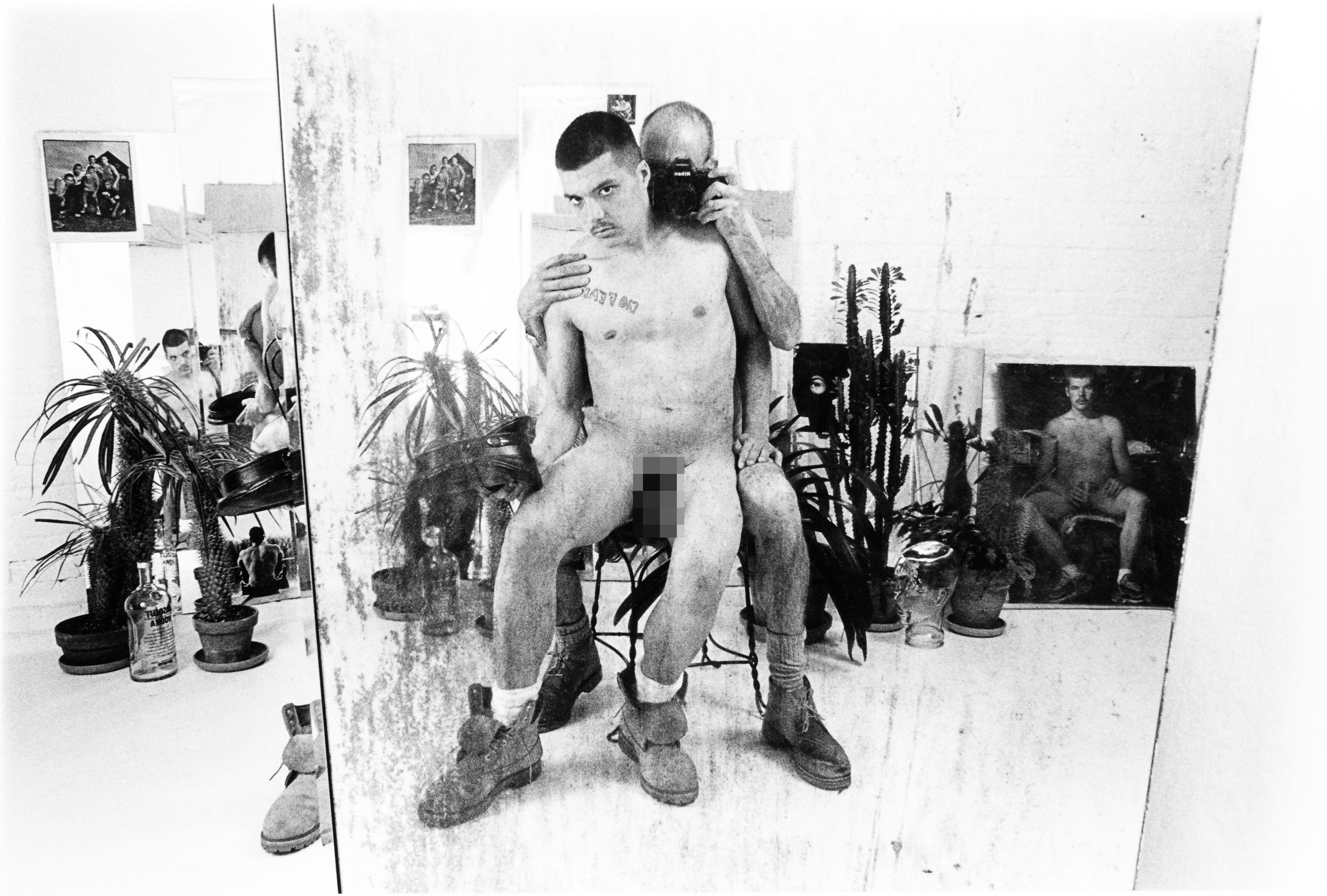 Black and white image showing two nude men embracing in an artist's studio with plants, artworks on walls, and scattered materials.