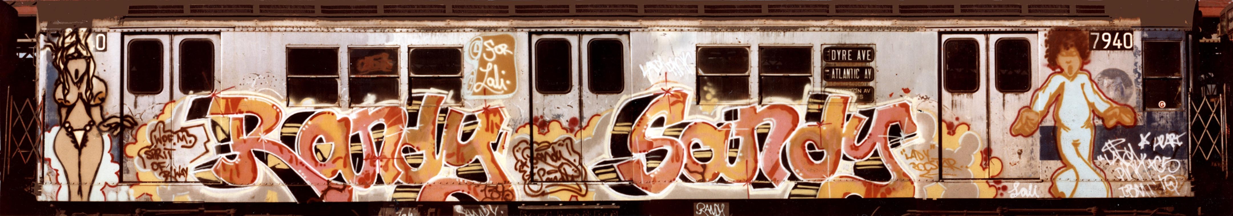 White train carriage with orange and brown graffiti lettering across the side, multiple rectangular windows above the text.