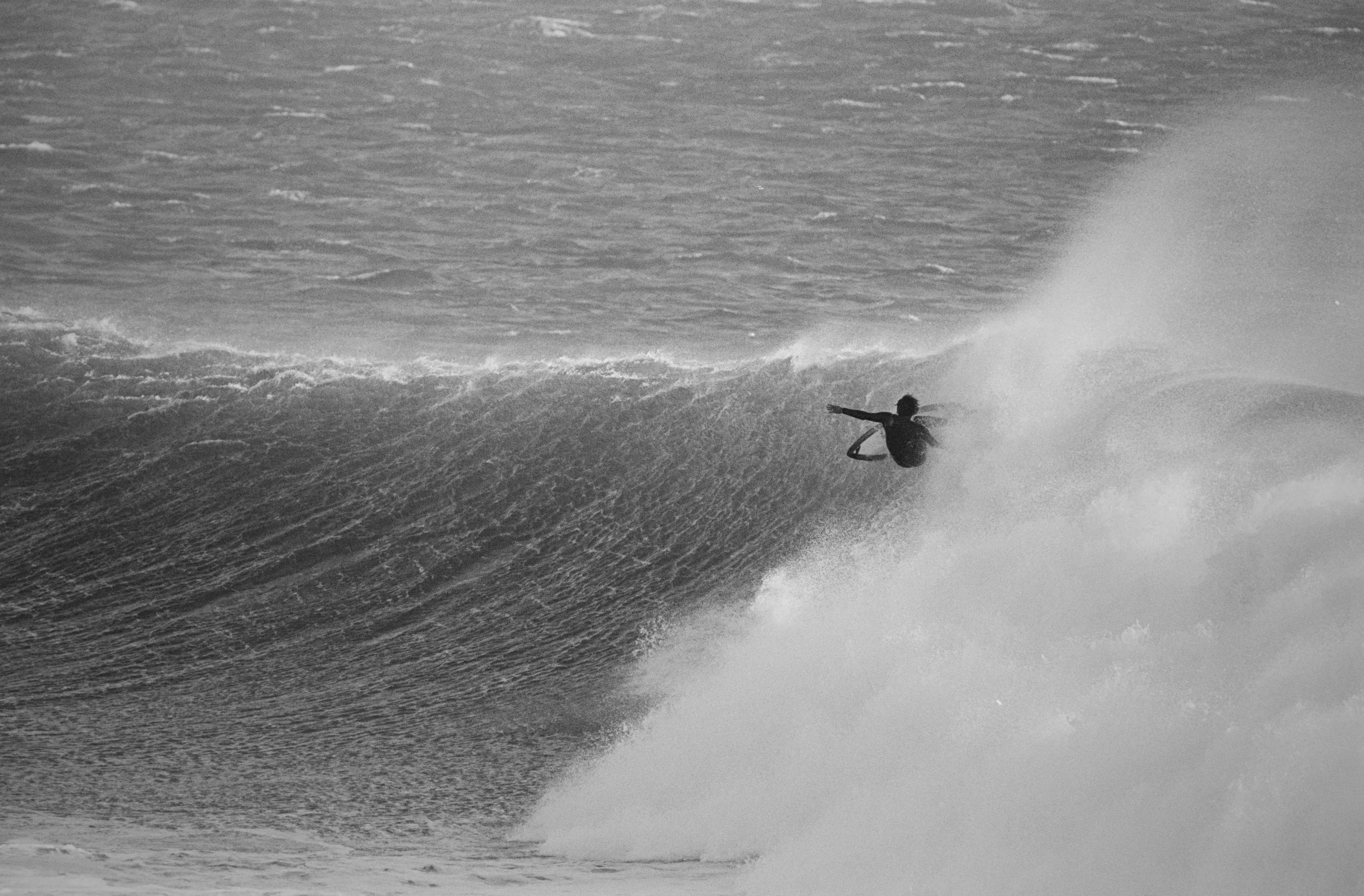 Surfer riding a large wave with spray and mist, black and white image showing ocean texture and water movement.