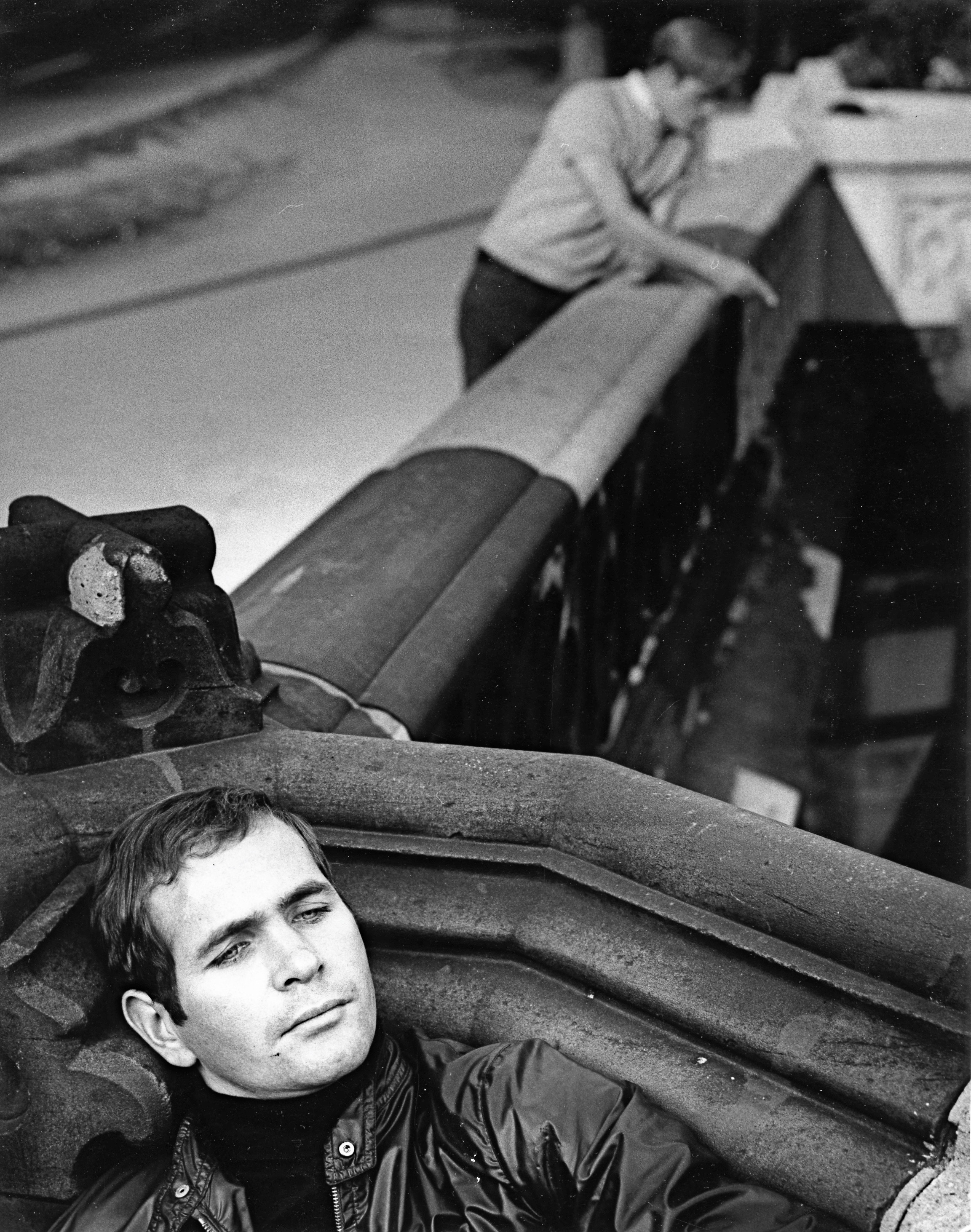 Black and white image showing man in leather jacket lying amongst large tyres in foreground, with another person working on structure in background.
