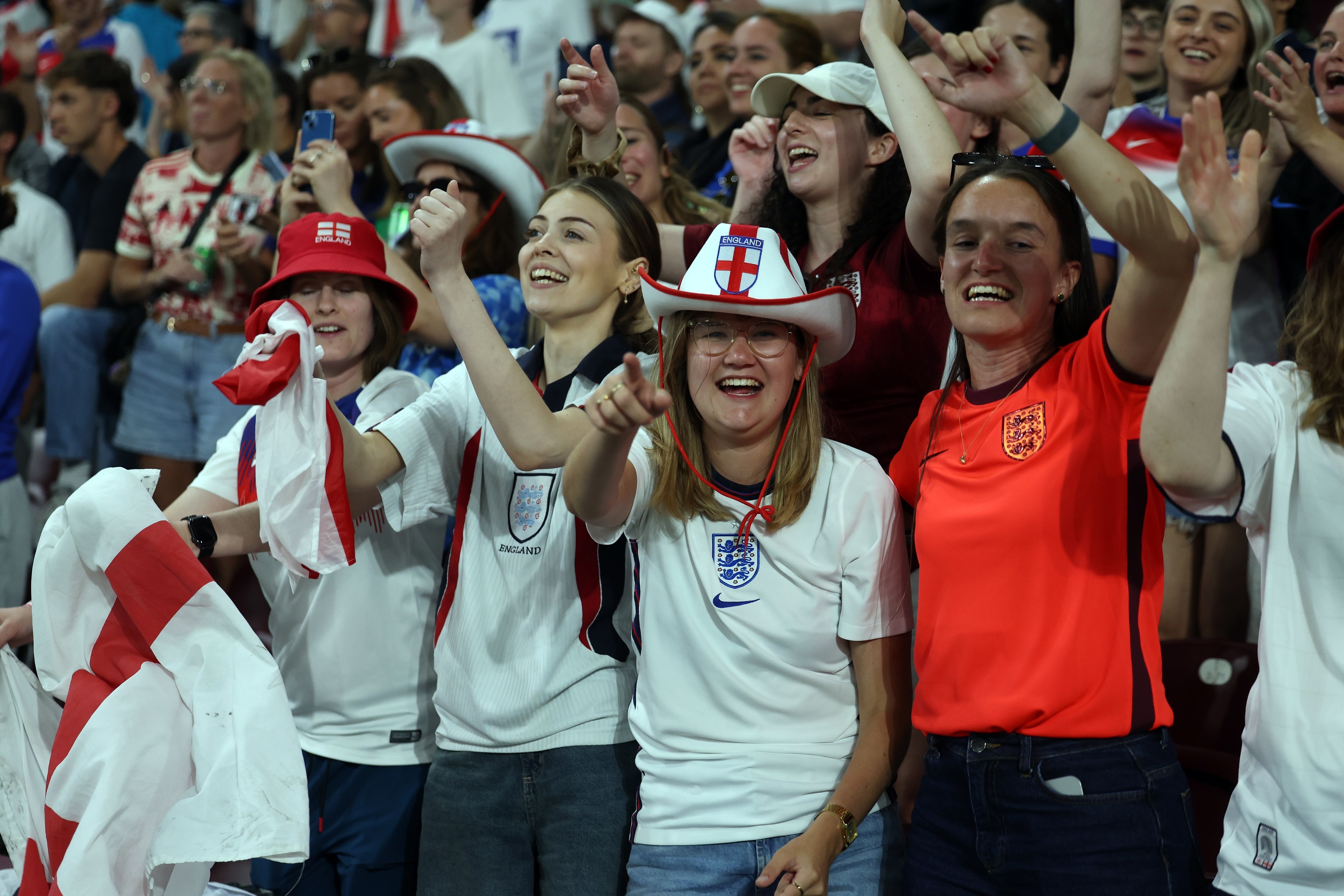 England football supporters celebrating in stadium stands wearing white jerseys, red and white hats, and face paint whilst cheering.