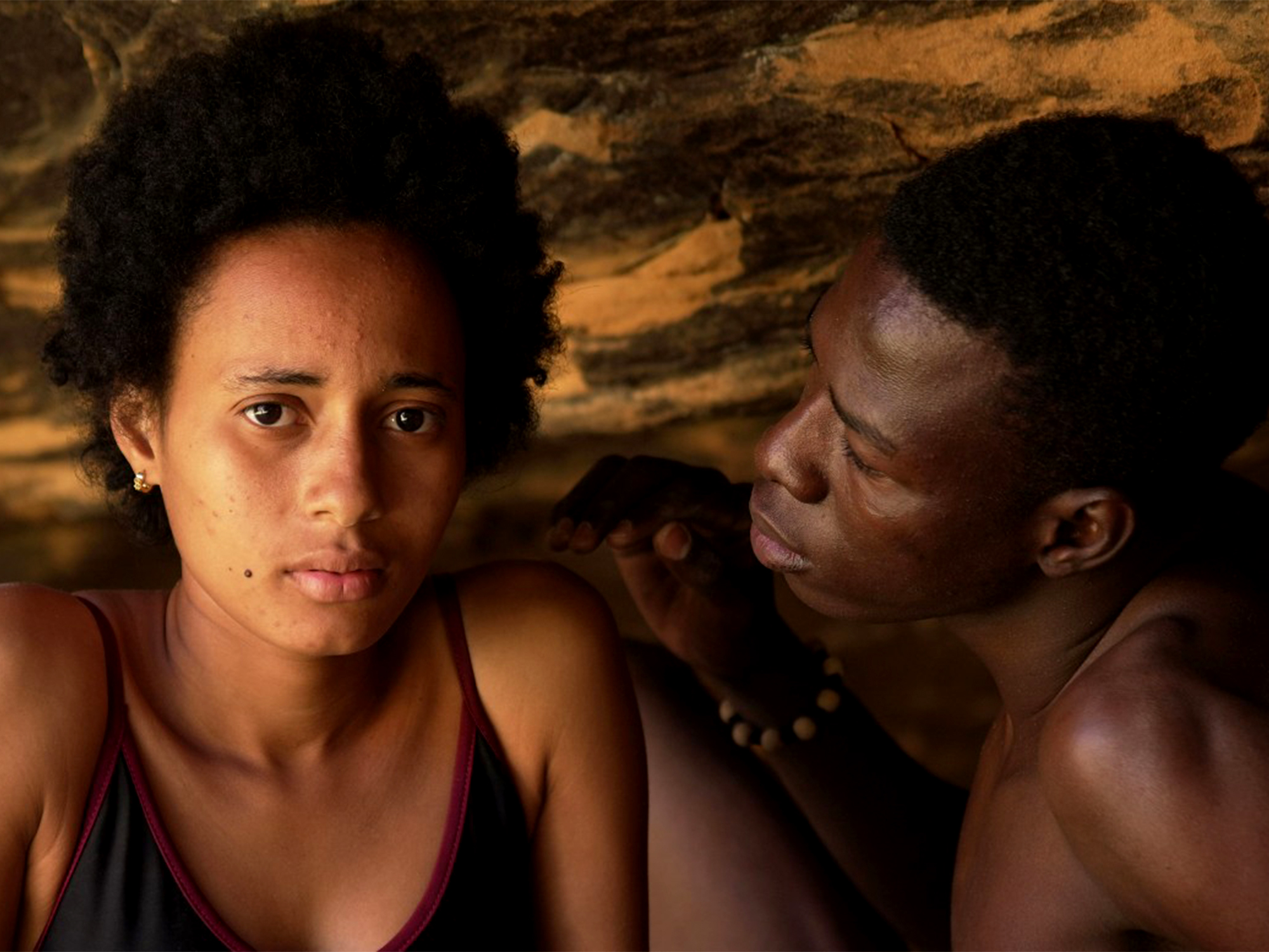 Two people lying close together against rough stone wall with warm orange and brown tones. Woman faces camera, man in profile beside her.