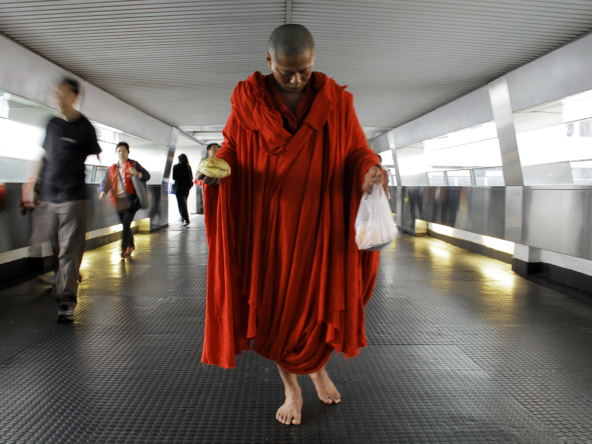 A Taiwanese man with a shaved head carrying a bright orange robe walking in a tunnel, carrying a sandwich in one hand and a plastic bag in the other, with other people walking in the background.