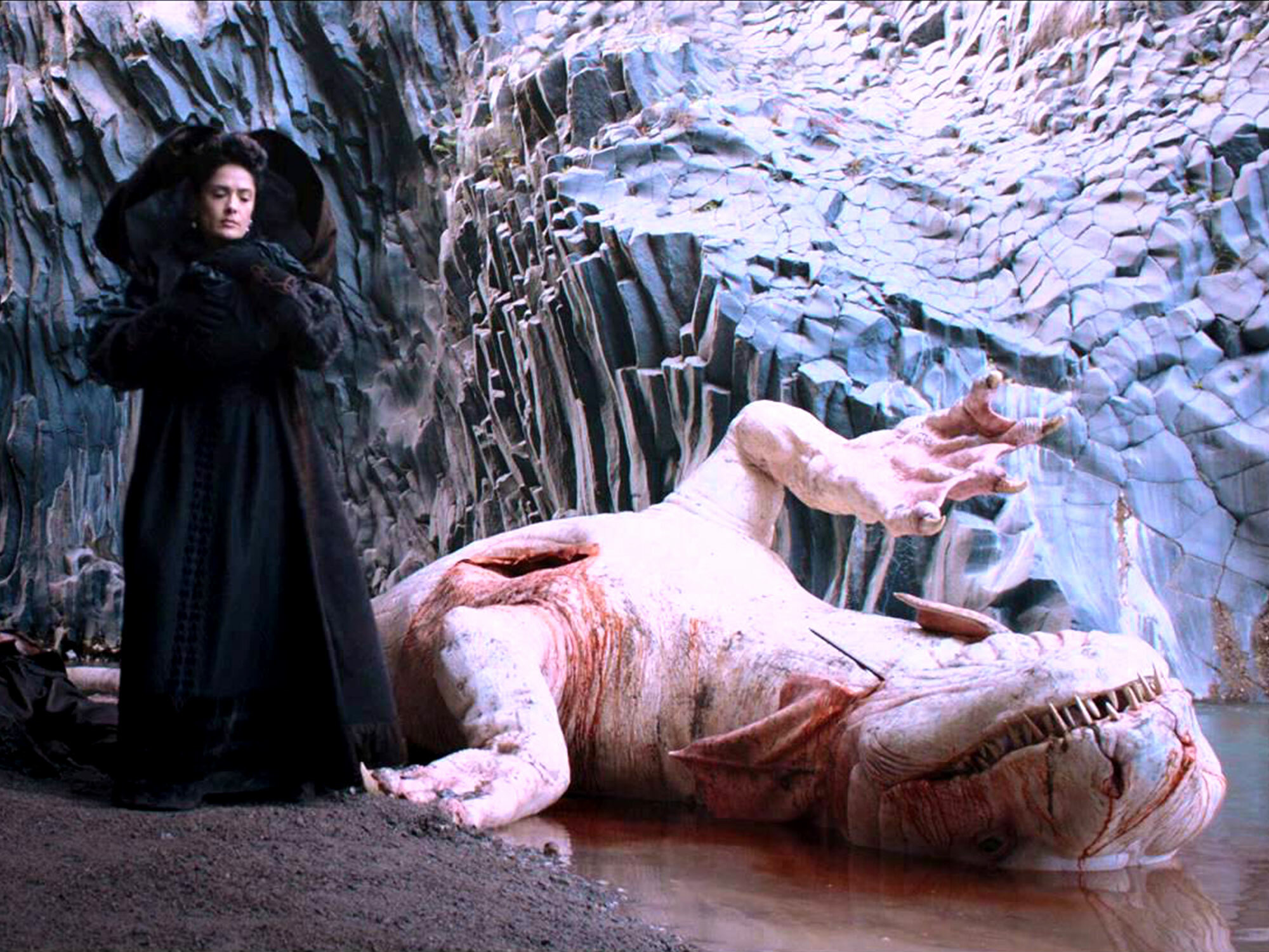 A hooded figure stands in a cavern of jagged ice formations, overlooking a reclining, partially disrobed figure lying on the ground.