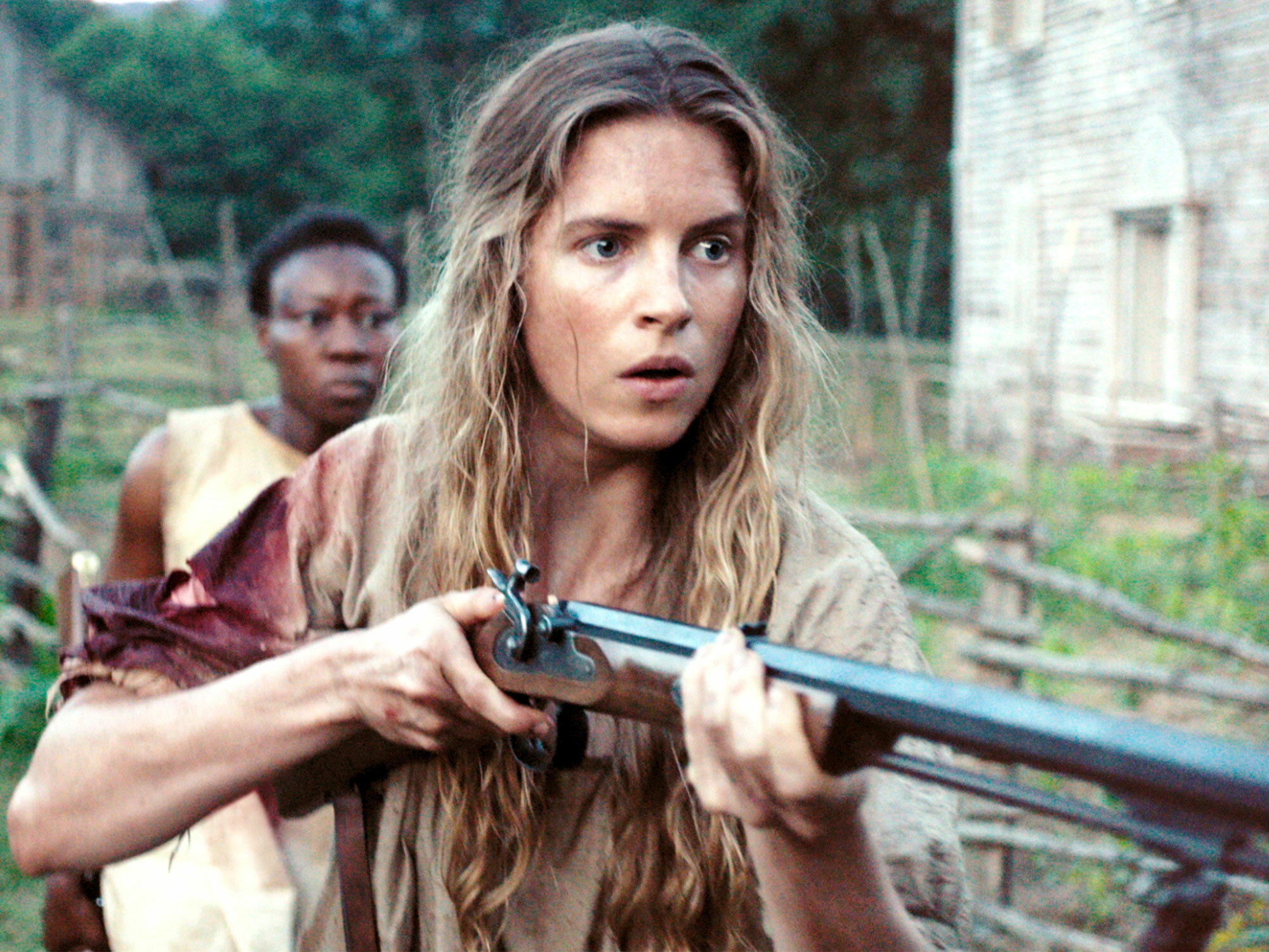 A woman with long, wavy hair holds a rifle, with a man in the background.