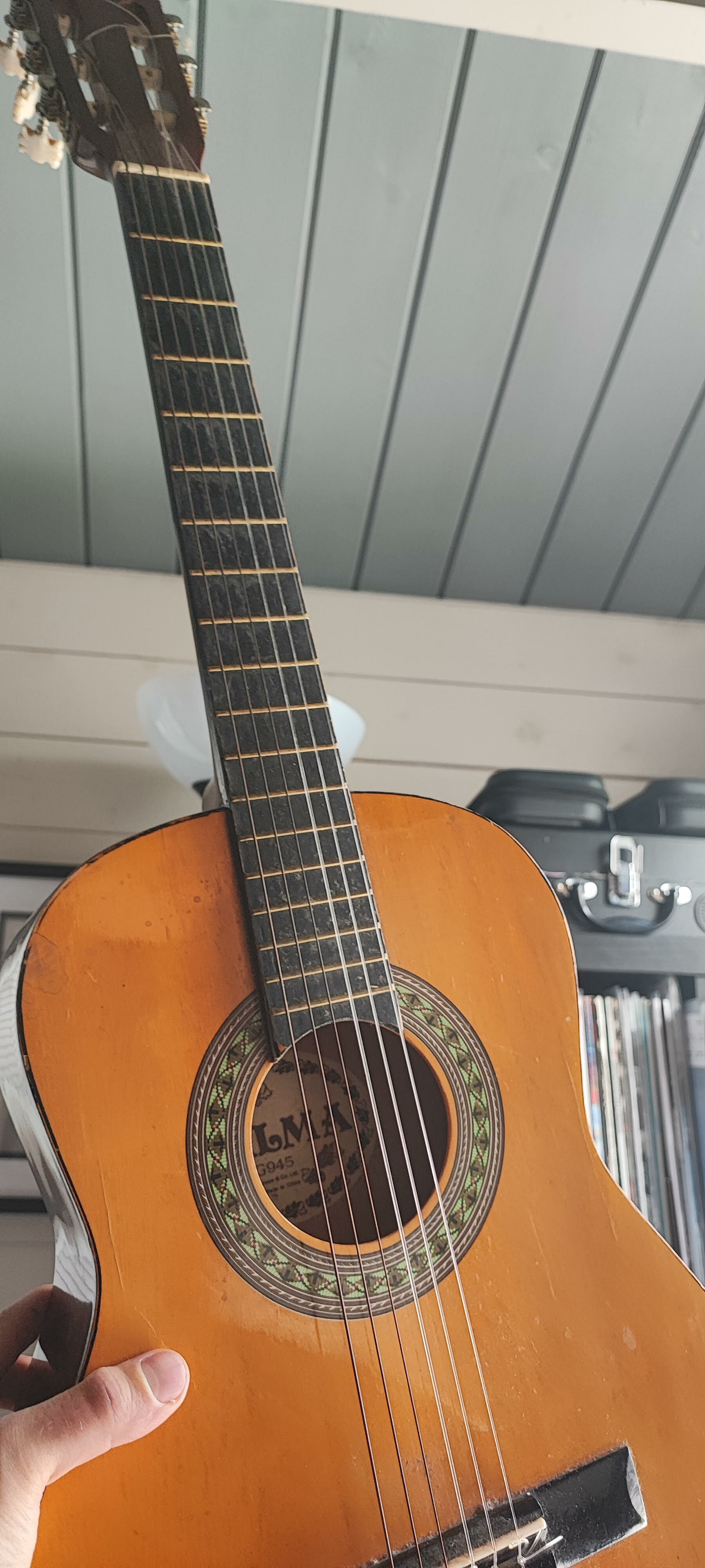 Classical guitar with natural wood body and dark fretboard being held by hand, with white ceiling and shelving visible in background.