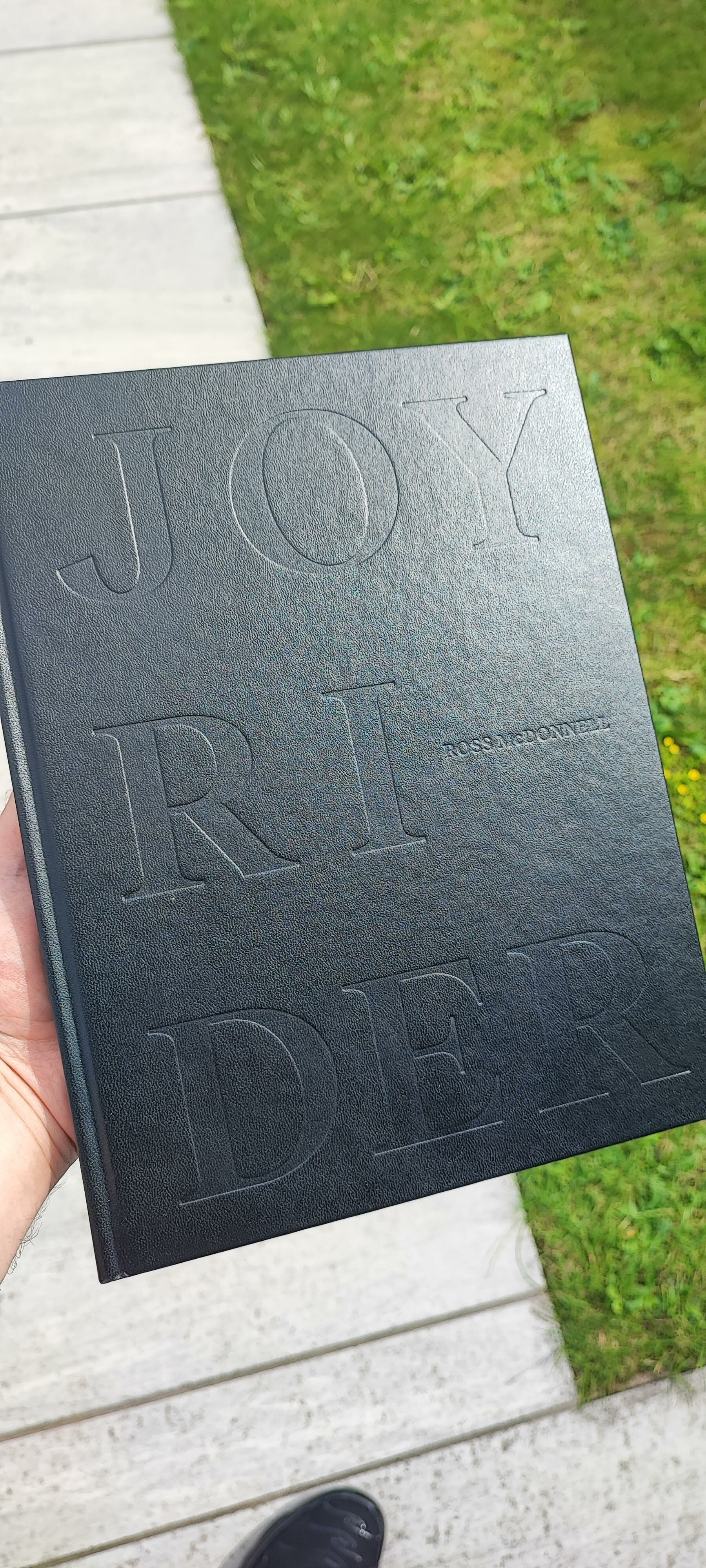 Hand holding dark grey book with embossed text "JOY" and "RIDE" visible, green grass and concrete pavement in background.