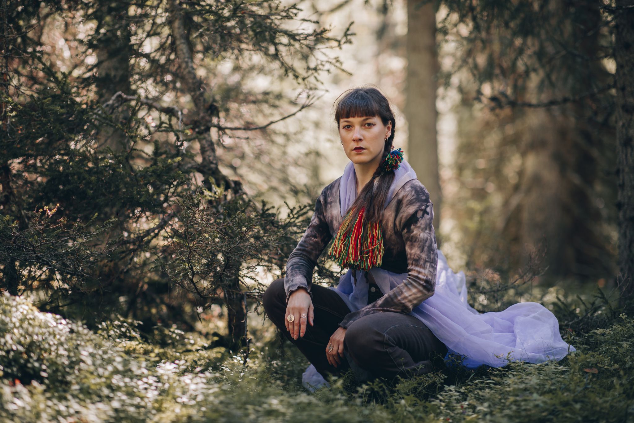 Woman in traditional Korean hanbok with grey jacket and white skirt sits amongst trees with dappled sunlight filtering through branches.