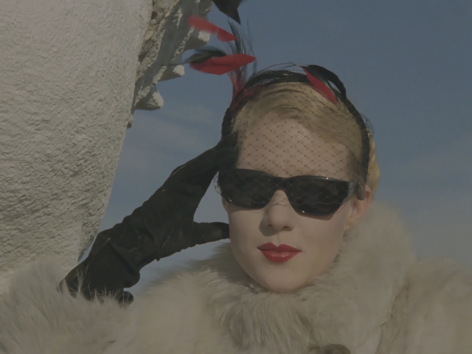 A blonde woman wearing black sunglasses, a headscarf, black gloves and a white fur coat.