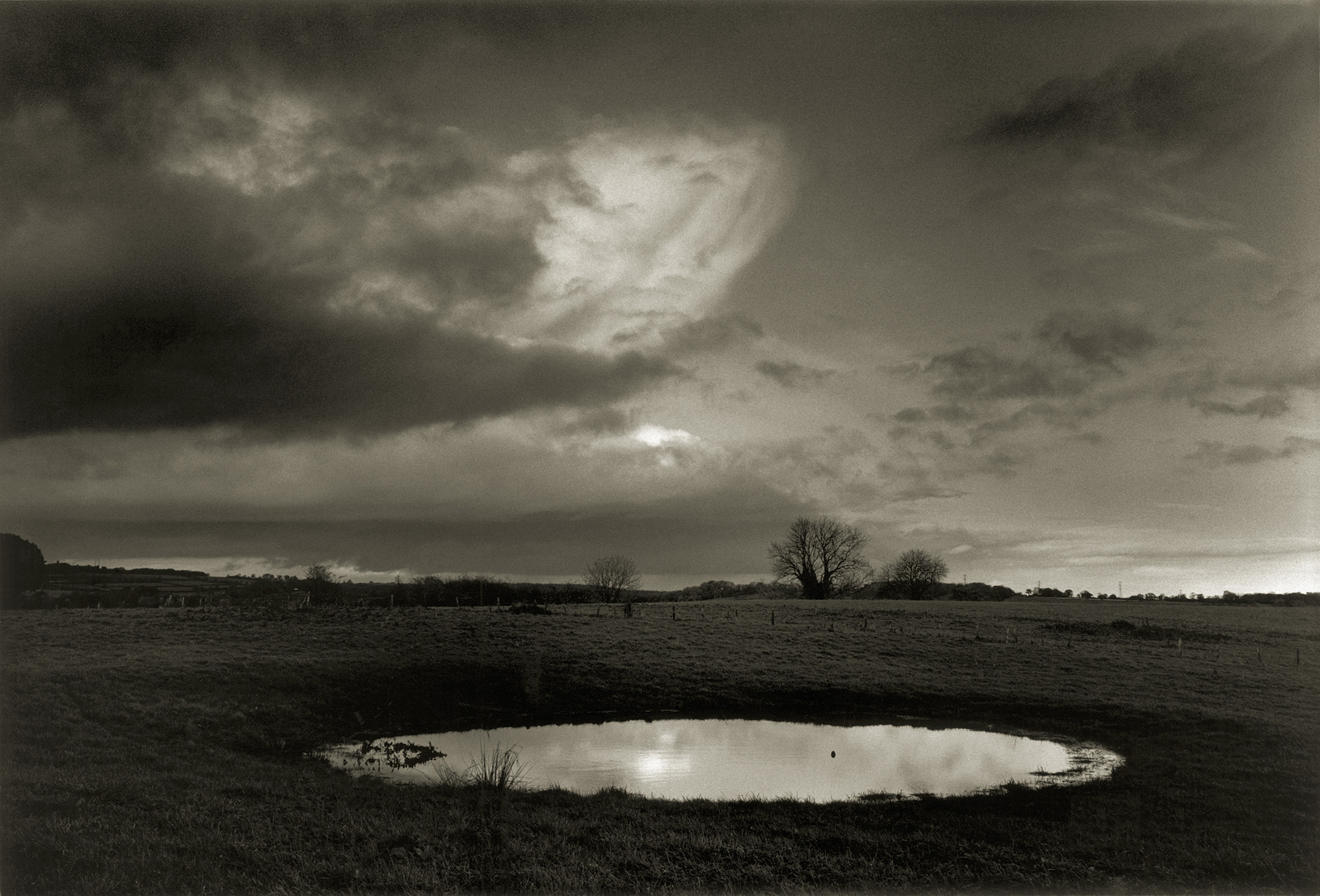 Dramatic cloudy sky over rural landscape with small pond reflecting light, bare trees on horizon, muted sepia tones throughout.