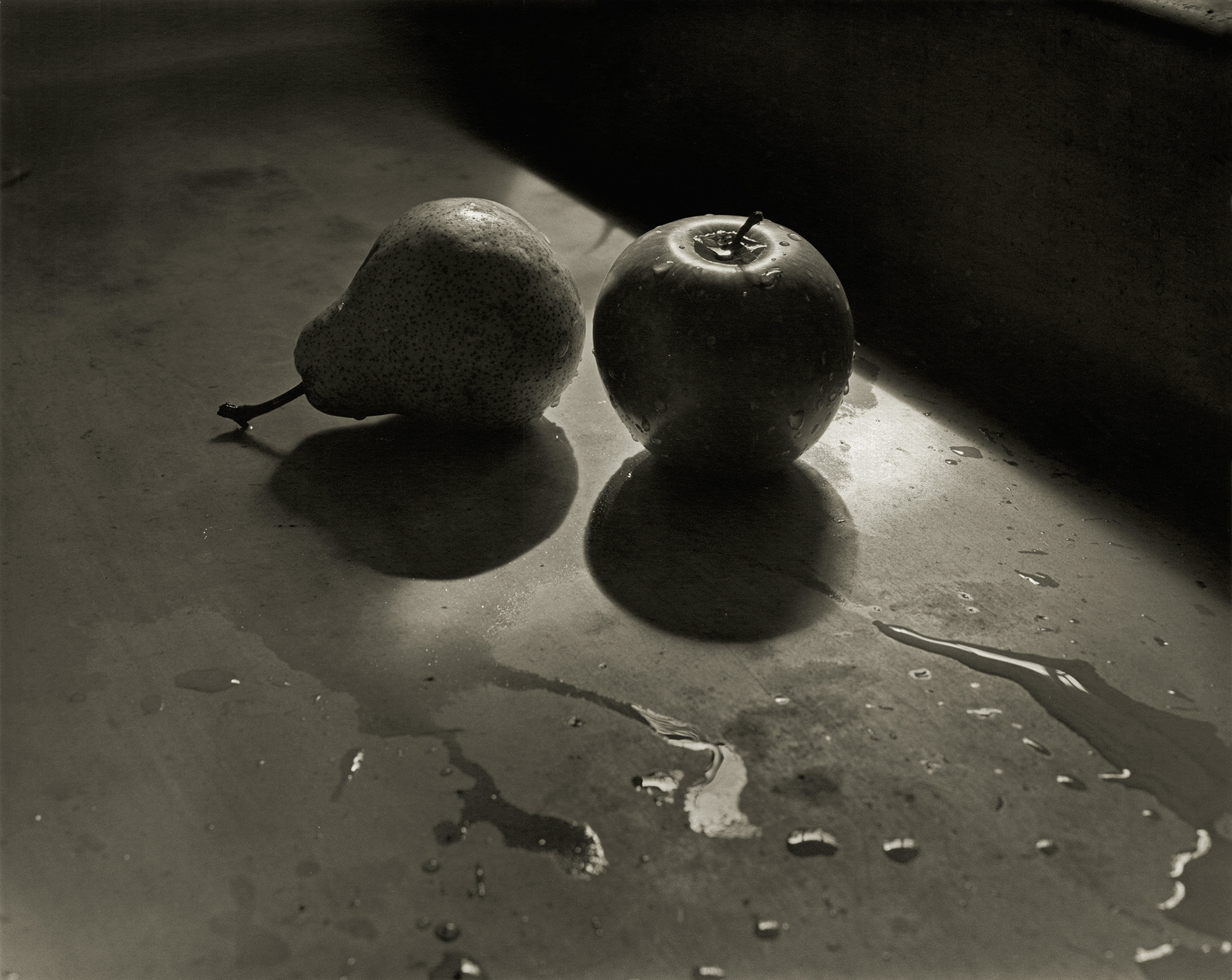 Two apples on wet surface with reflections in dramatic lighting, dark shadows surrounding illuminated fruit in centre.